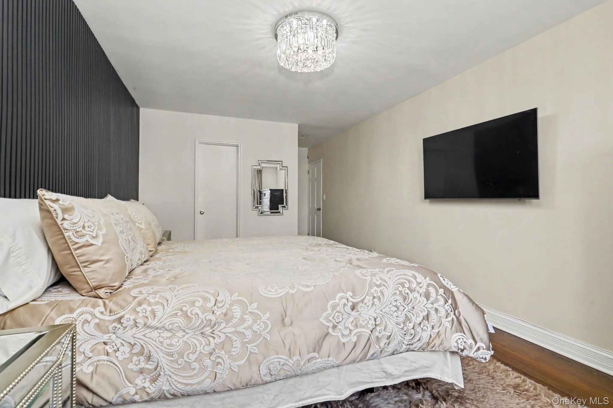 Textured accent wall with vertical paneling, mounted television, wood-finish flooring, and a crystal chandelier Textured accent wall with vertical paneling, mounted television, wood-finish flooring, and a crystal chandelier