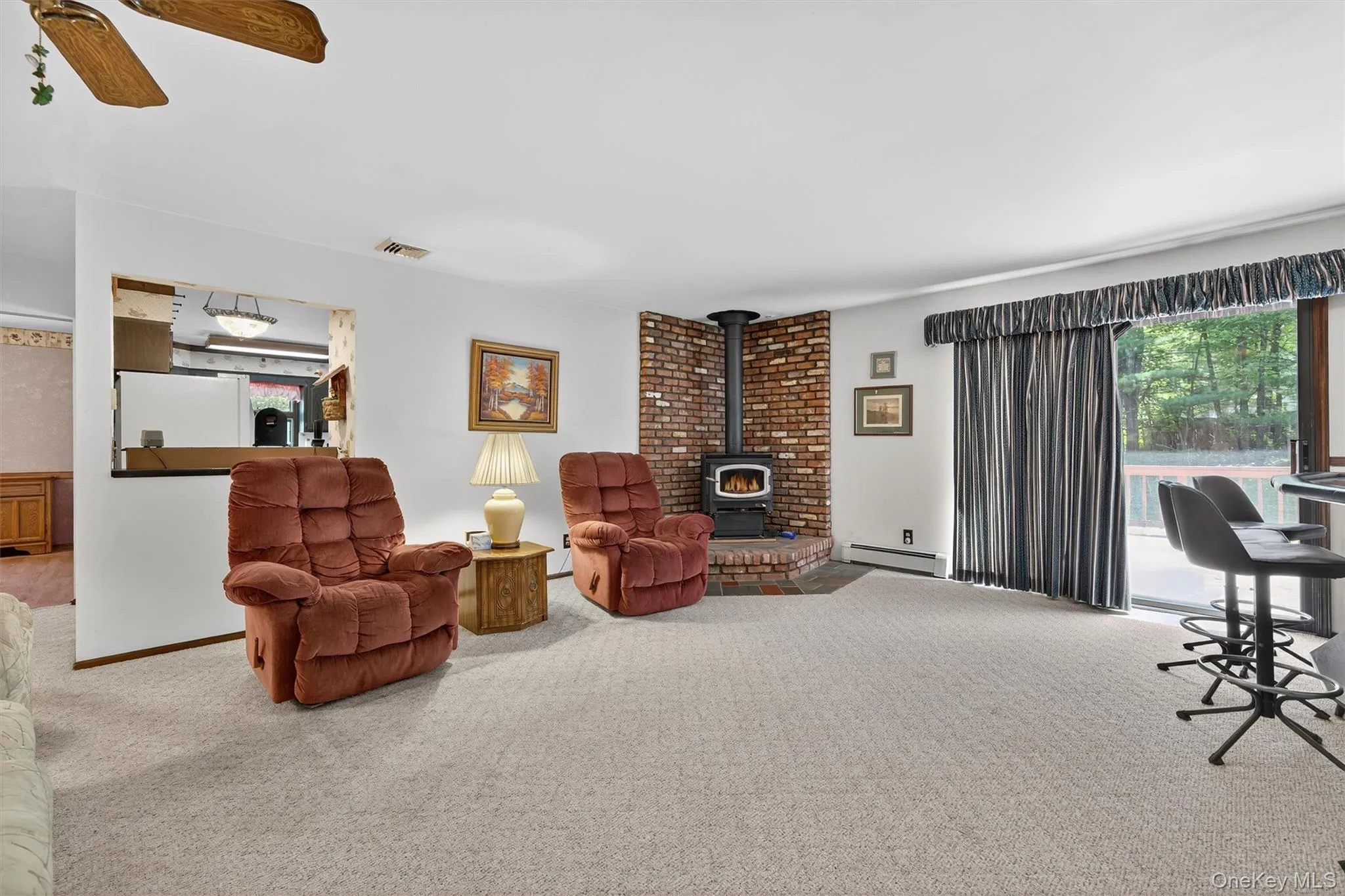 Carpeted living room with a baseboard heating unit, a wood stove, and ceiling fan Carpeted living room with a baseboard heating unit, a wood stove, and ceiling fan