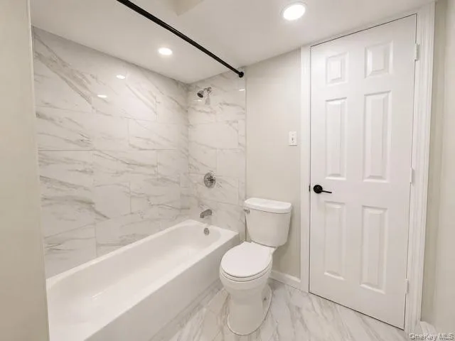 White porcelain bathtub with chrome fixtures, large format wall tile, recessed ceiling lighting, a standard toilet, and a paneled door with a matte black lever handle White porcelain bathtub with chrome fixtures, large format wall tile, recessed ceiling lighting, a standard toilet, and a paneled door with a matte black lever handle