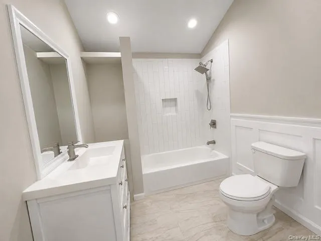 Modern bathroom featuring a white vanity with integrated sink, rectangular framed mirror, built-in tub with vertical tile surround, and wainscoting detail Modern bathroom featuring a white vanity with integrated sink, rectangular framed mirror, built-in tub with vertical tile surround, and wainscoting detail