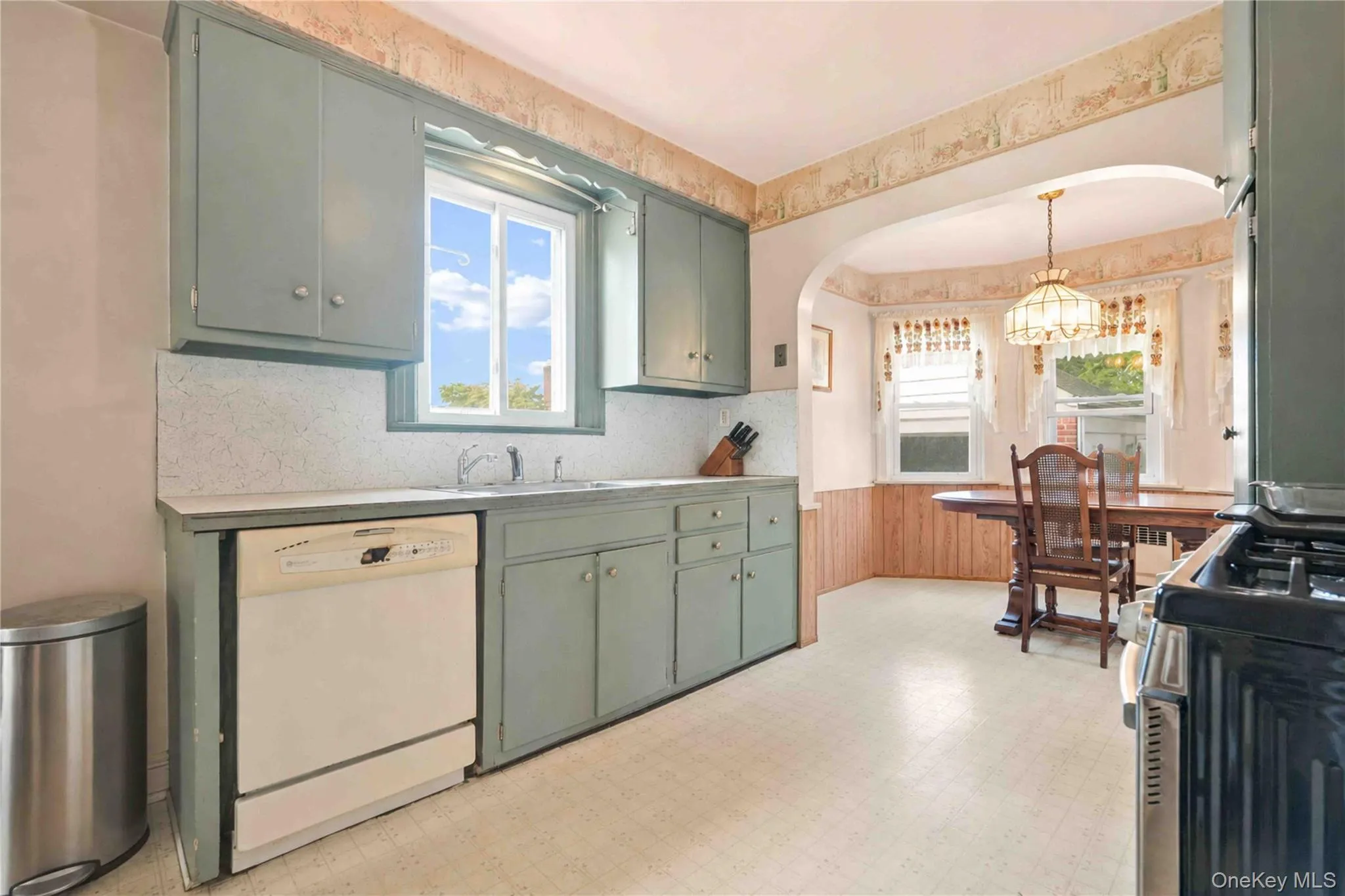 Galley kitchen featuring sage-green cabinetry, stainless steel appliances, wood-finish countertops, a window with natural light, and wood-finish flooring that extends into the adjoining dining area with original architectural details and pendant lighting Galley kitchen featuring sage-green cabinetry, stainless steel appliances, wood-finish countertops, a window with natural light, and wood-finish flooring that extends into the adjoining dining area with original architectural details and pendant lighting