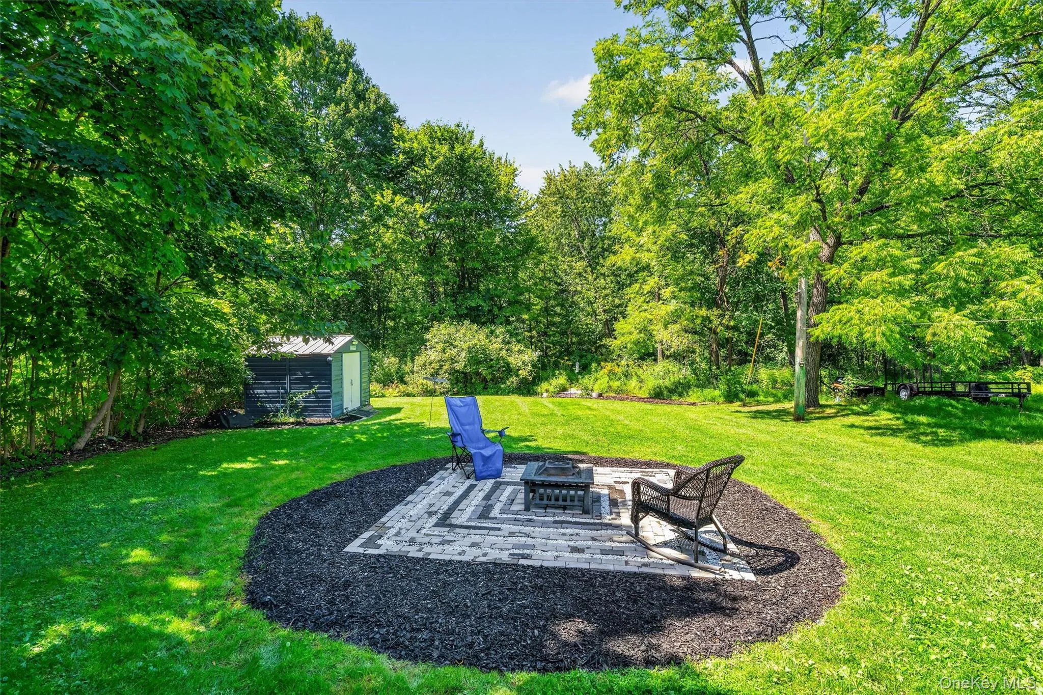 Expansive lawn featuring a paver patio with a fire pit, surrounded by mature trees Expansive lawn featuring a paver patio with a fire pit, surrounded by mature trees