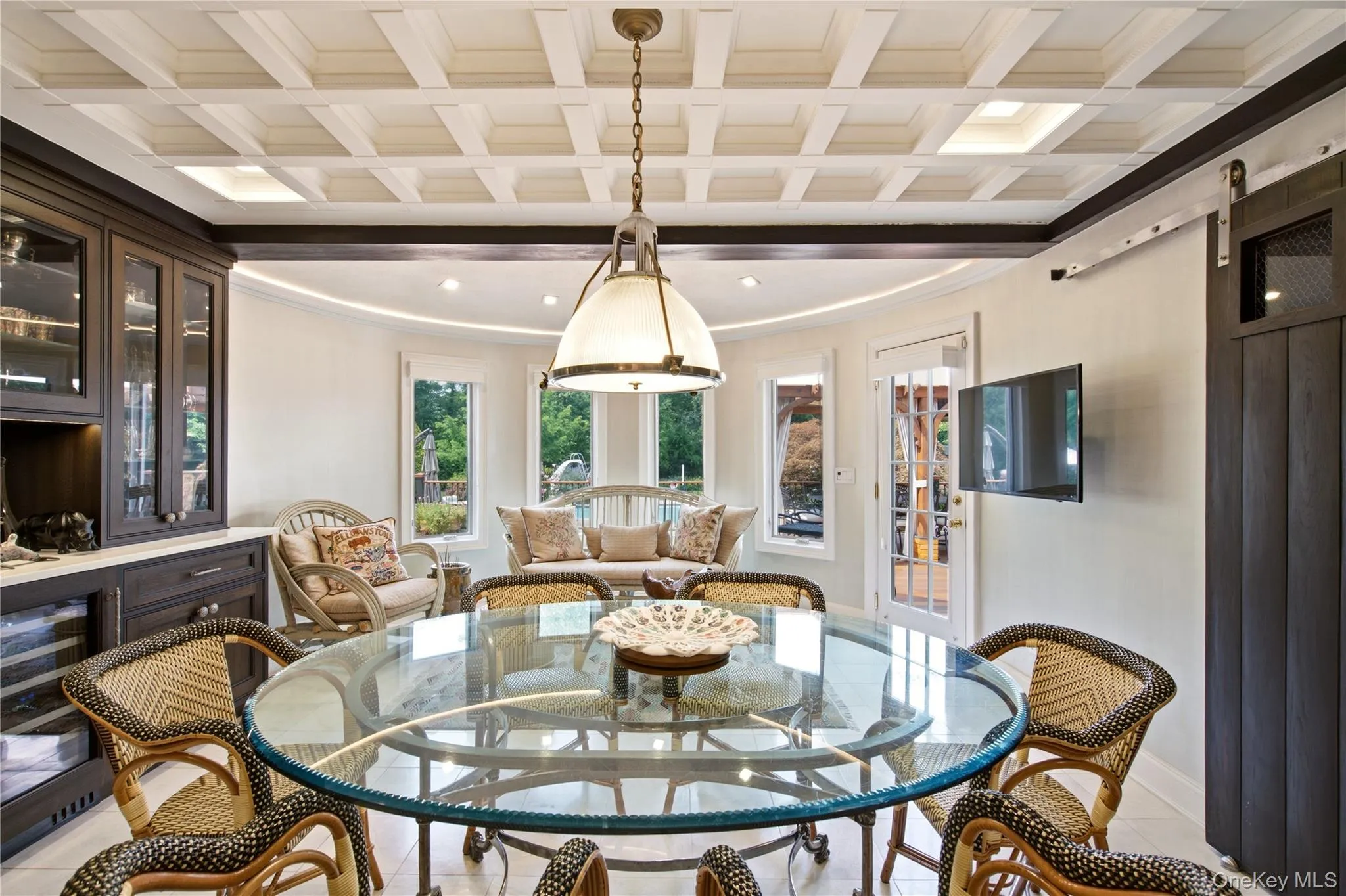 Recessed lighting illuminating a coffered ceiling, with glass-front cabinetry, a glass-top dining table, a white French door, and a dark wood-finish barn door Recessed lighting illuminating a coffered ceiling, with glass-front cabinetry, a glass-top dining table, a white French door, and a dark wood-finish barn door