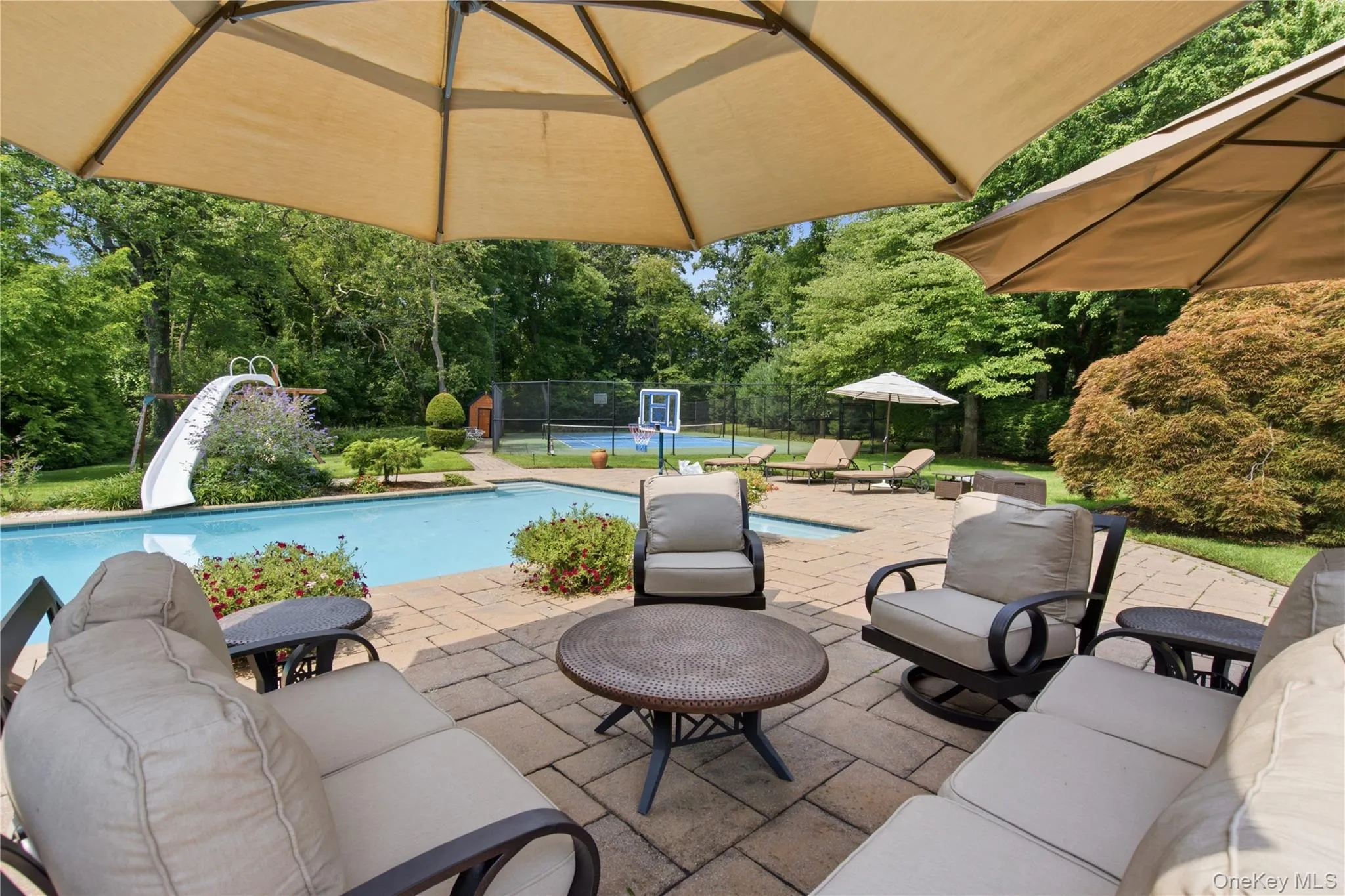 Paver patio with multiple shade umbrellas, a swimming pool with an attached slide, and a tennis court Paver patio with multiple shade umbrellas, a swimming pool with an attached slide, and a tennis court