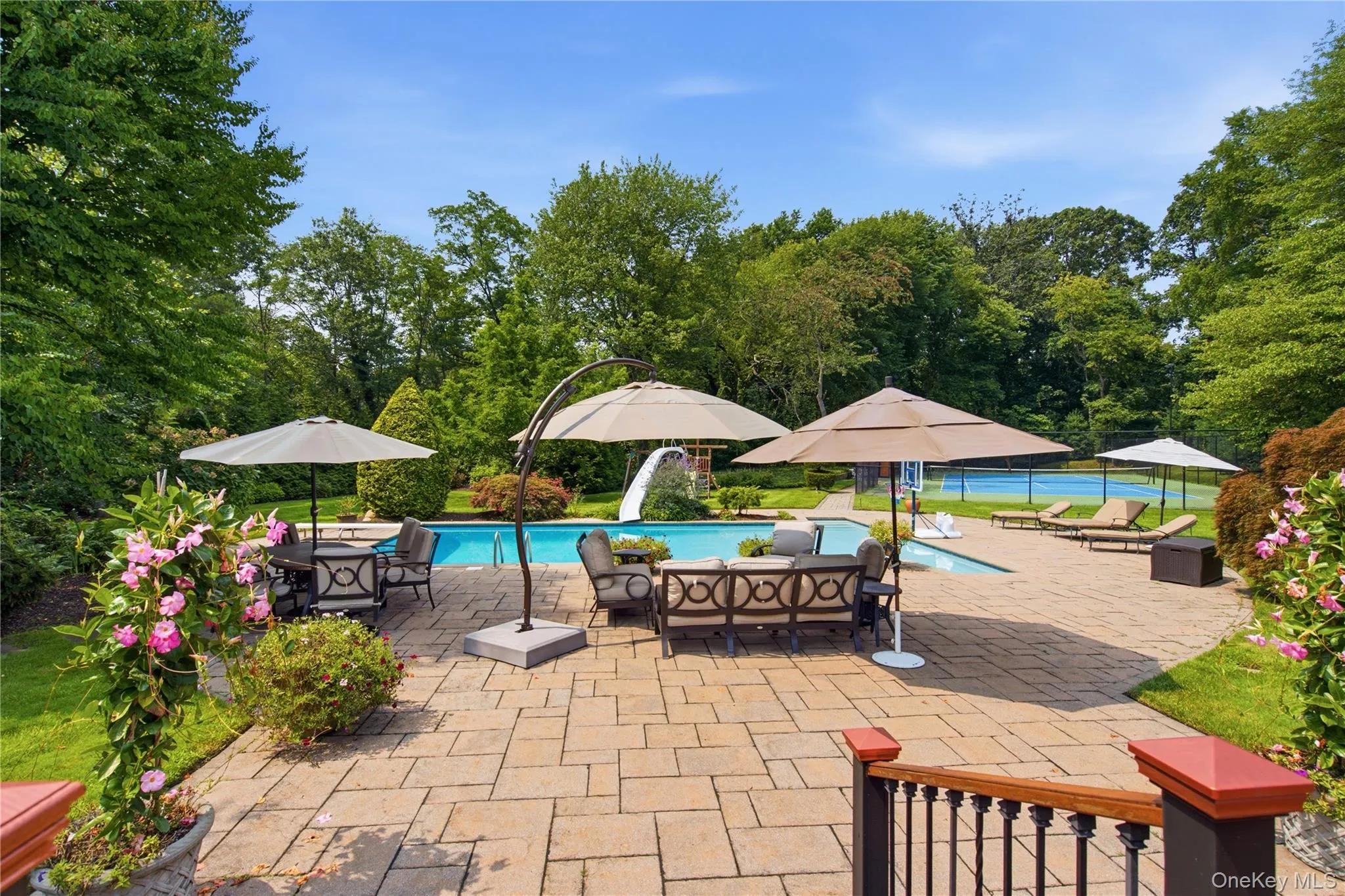 Expansive paver patio with an in-ground swimming pool, mature perimeter landscaping, and a fenced tennis court Expansive paver patio with an in-ground swimming pool, mature perimeter landscaping, and a fenced tennis court