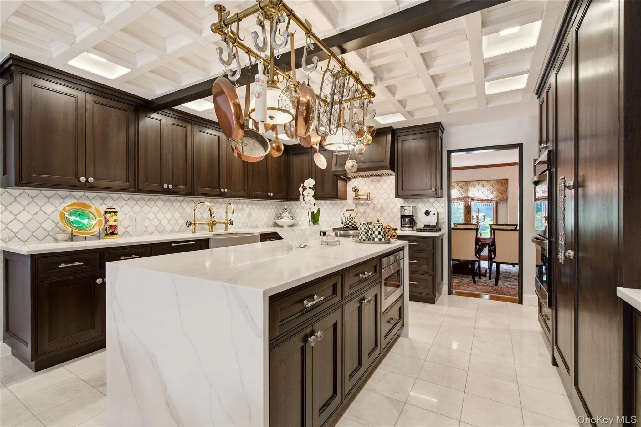 Black oak wood cabinetry, a large center island with a waterfall countertop, tiled backsplash, coffered ceiling, and integrated stainless steel double ovens Black oak wood cabinetry, a large center island with a waterfall countertop, tiled backsplash, coffered ceiling, and integrated stainless steel double ovens