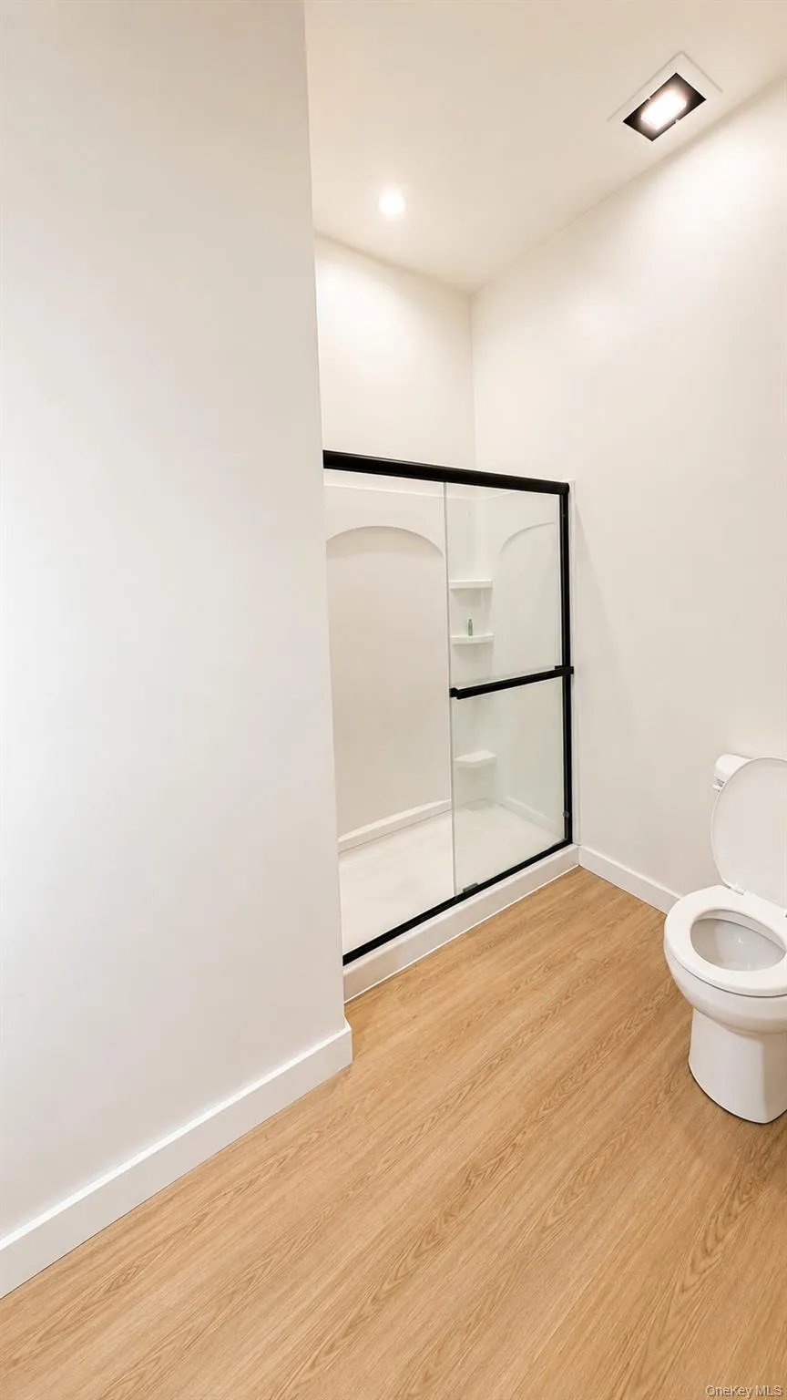 Walk-in shower with clear glass enclosure and black framing, white toilet, wood-finish flooring, recessed lighting Walk-in shower with clear glass enclosure and black framing, white toilet, wood-finish flooring, recessed lighting