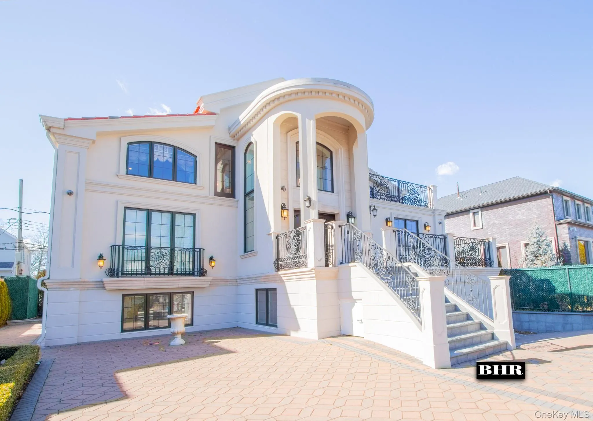 Stucco exterior featuring a grand curved entrance with columns, decorative iron railings, arched windows, and a paved brick driveway Stucco exterior featuring a grand curved entrance with columns, decorative iron railings, arched windows, and a paved brick driveway