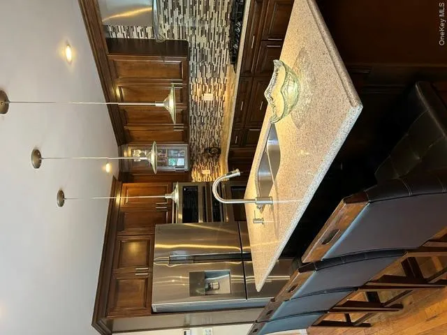 Gourmet kitchen featuring stainless steel appliances, dark wood cabinetry, a mosaic tile backsplash, a kitchen island with a double basin sink, and pendant lighting Gourmet kitchen featuring stainless steel appliances, dark wood cabinetry, a mosaic tile backsplash, a kitchen island with a double basin sink, and pendant lighting