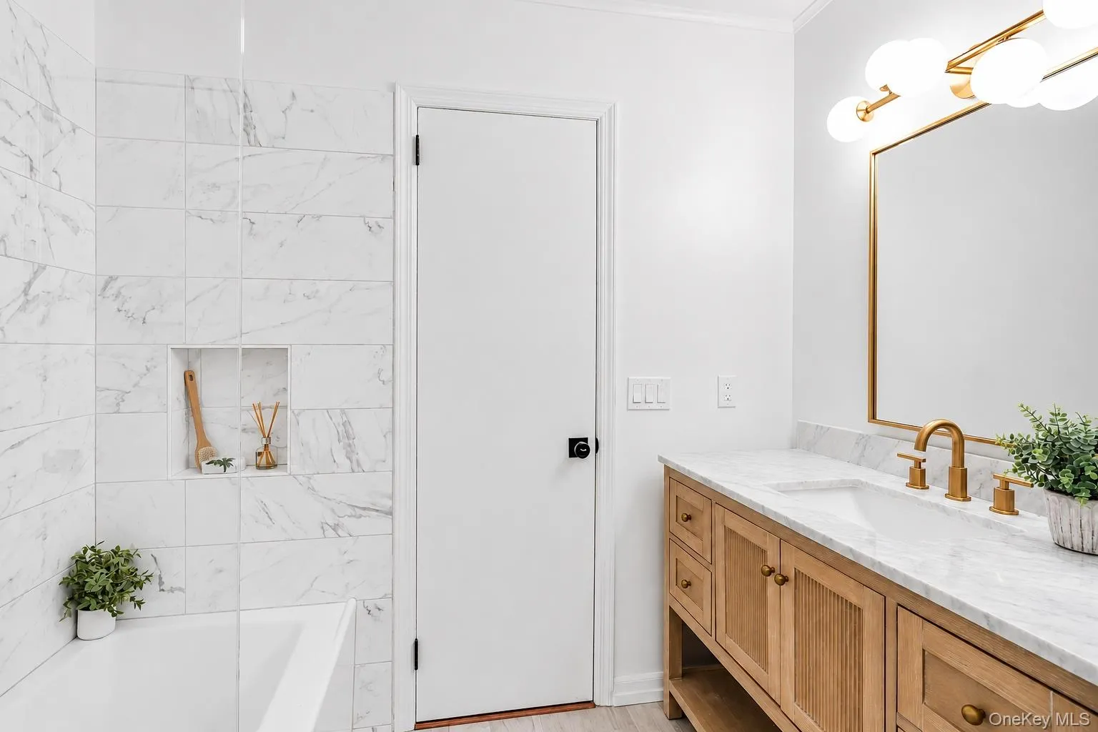 Bathroom featuring a natural wood-finish vanity with a white countertop, gold-toned faucet and cabinet hardware, and a framed mirror with a modern light fixture Bathroom featuring a natural wood-finish vanity with a white countertop, gold-toned faucet and cabinet hardware, and a framed mirror with a modern light fixture