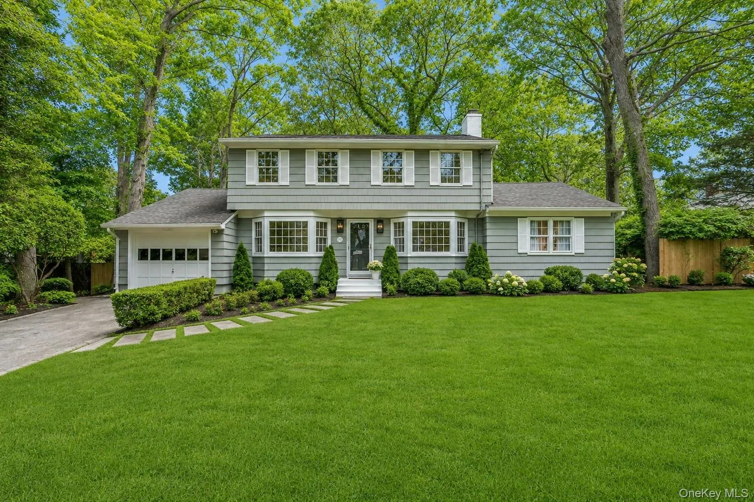 Two-story facade with gray wood, white window shutters, a prominent bay window, attached garage, and a lush front lawn with mature trees Two-story facade with gray wood, white window shutters, a prominent bay window, attached garage, and a lush front lawn with mature trees