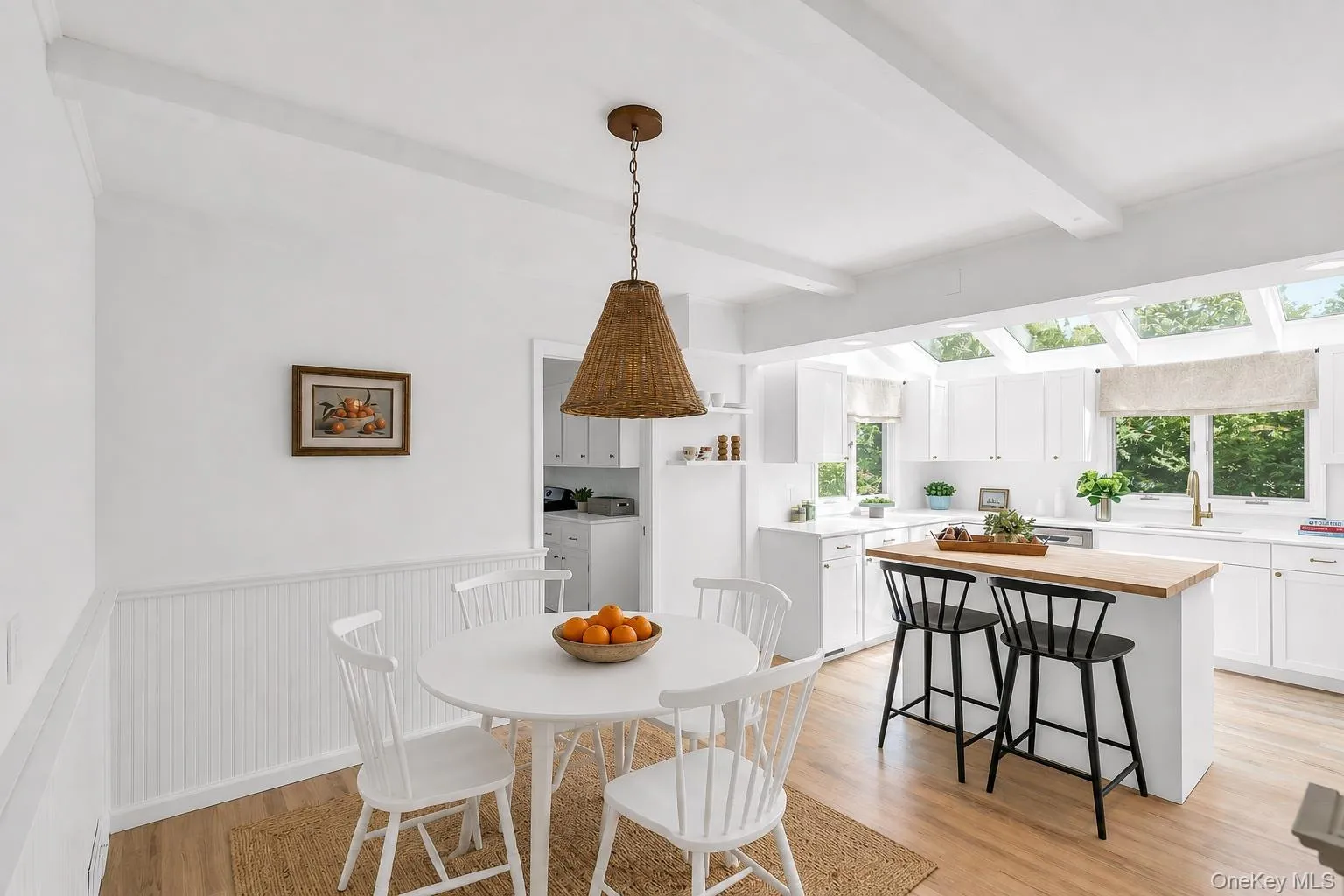 White shaker cabinetry, light wood-finish flooring, exposed ceiling beams, a central island with wood butcher block countertop, and multiple skylights White shaker cabinetry, light wood-finish flooring, exposed ceiling beams, a central island with wood butcher block countertop, and multiple skylights
