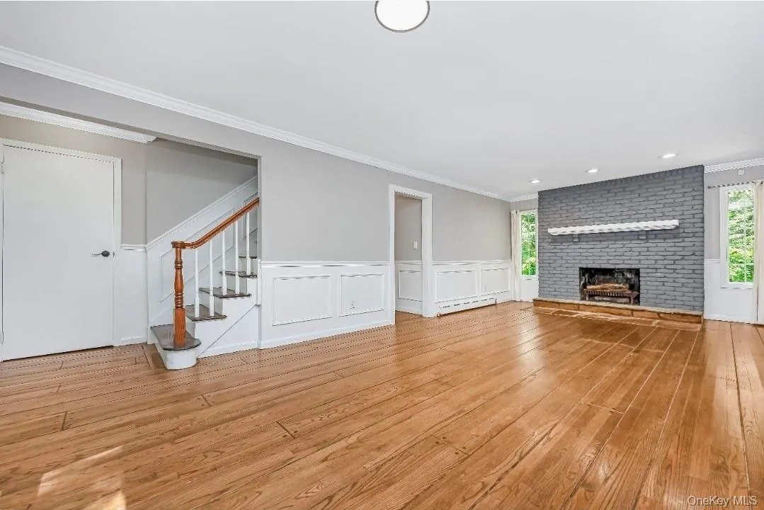 Expansive room featuring wood-finish flooring, a painted brick fireplace with a mantel, white wainscoting, and a staircase with a wood handrail Expansive room featuring wood-finish flooring, a painted brick fireplace with a mantel, white wainscoting, and a staircase with a wood handrail