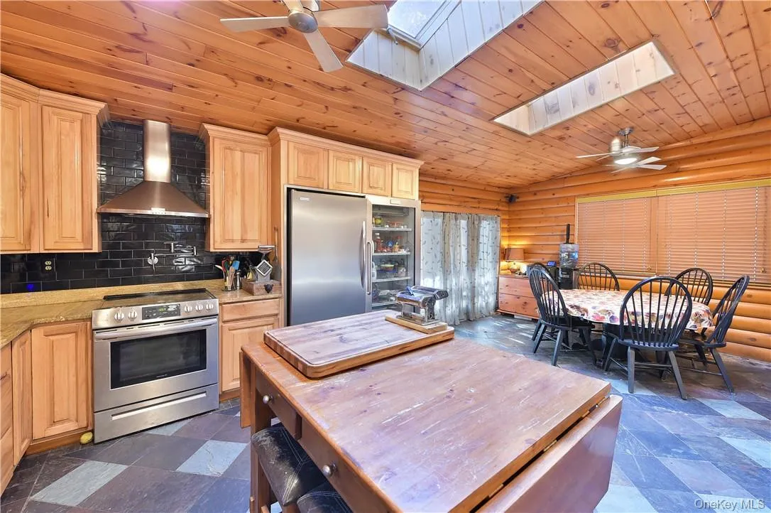 Kitchen featuring wood ceiling, wall chimney range hood, stainless steel appliances, light brown cabinetry, and ceiling fan Kitchen featuring wood ceiling, wall chimney range hood, stainless steel appliances, light brown cabinetry, and ceiling fan