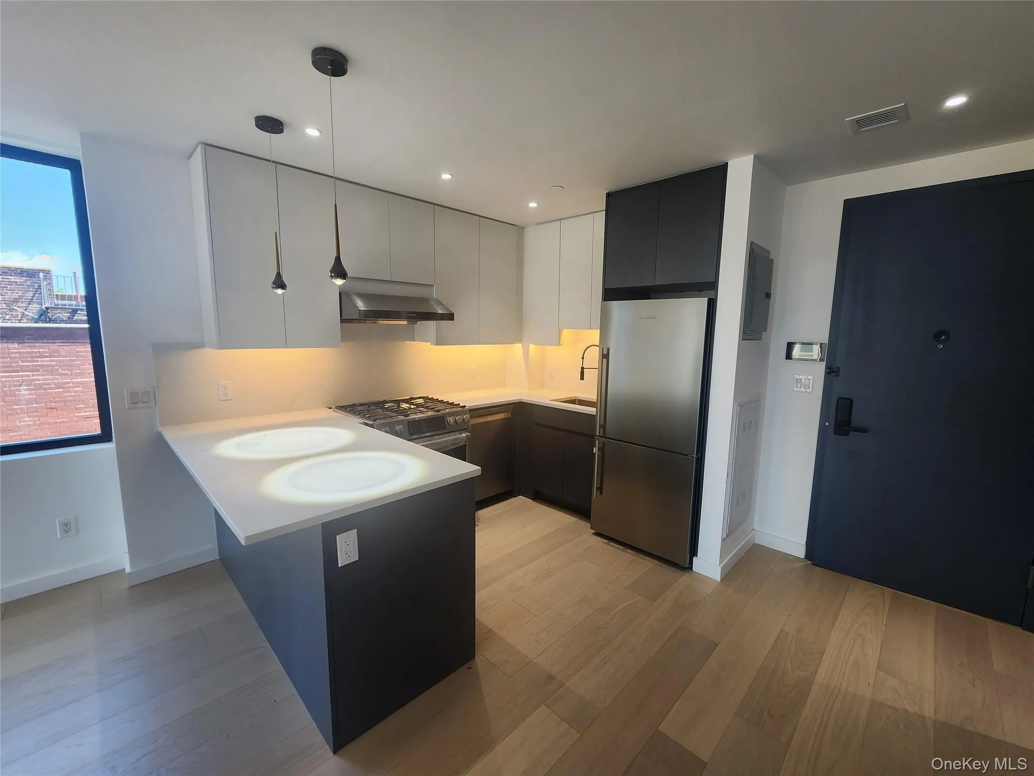 Modern kitchen featuring light wood-finish flooring, stainless steel appliances, dark lower cabinetry, light upper cabinetry, and a white countertop with an integrated breakfast bar Modern kitchen featuring light wood-finish flooring, stainless steel appliances, dark lower cabinetry, light upper cabinetry, and a white countertop with an integrated breakfast bar