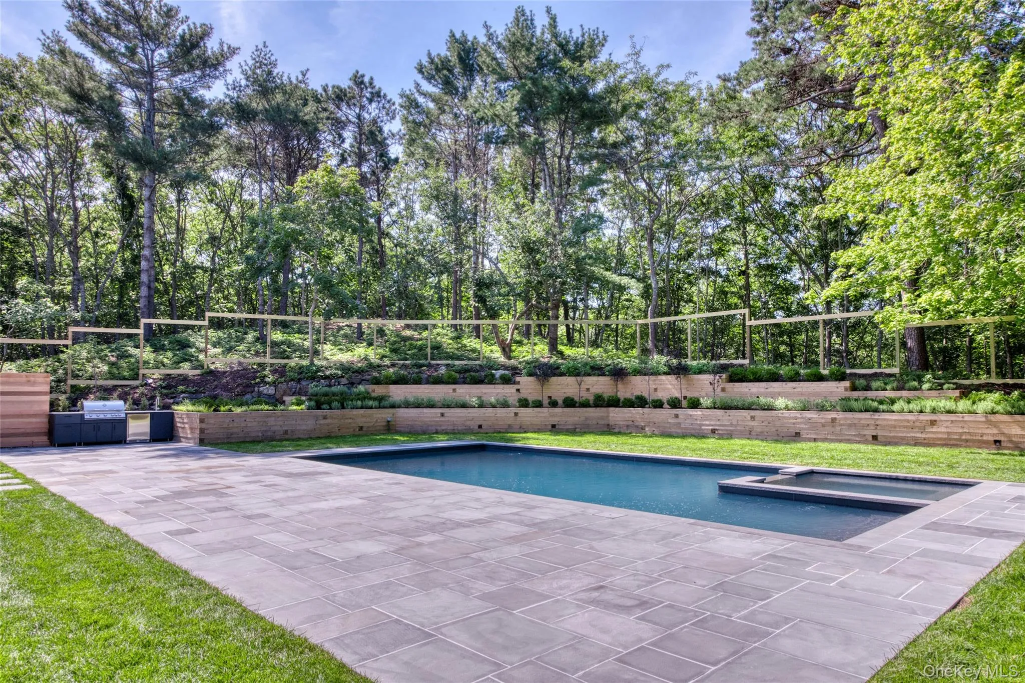 Swimming pool with integrated spa, expansive paver patio, built-in outdoor kitchen, tiered landscaping, and mature perimeter trees Swimming pool with integrated spa, expansive paver patio, built-in outdoor kitchen, tiered landscaping, and mature perimeter trees