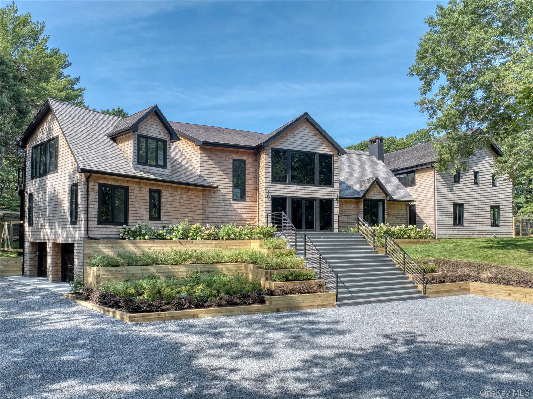 Shingle-style exterior with black trim windows, multi-level tiered landscaping, broad front steps, and a gravel driveway