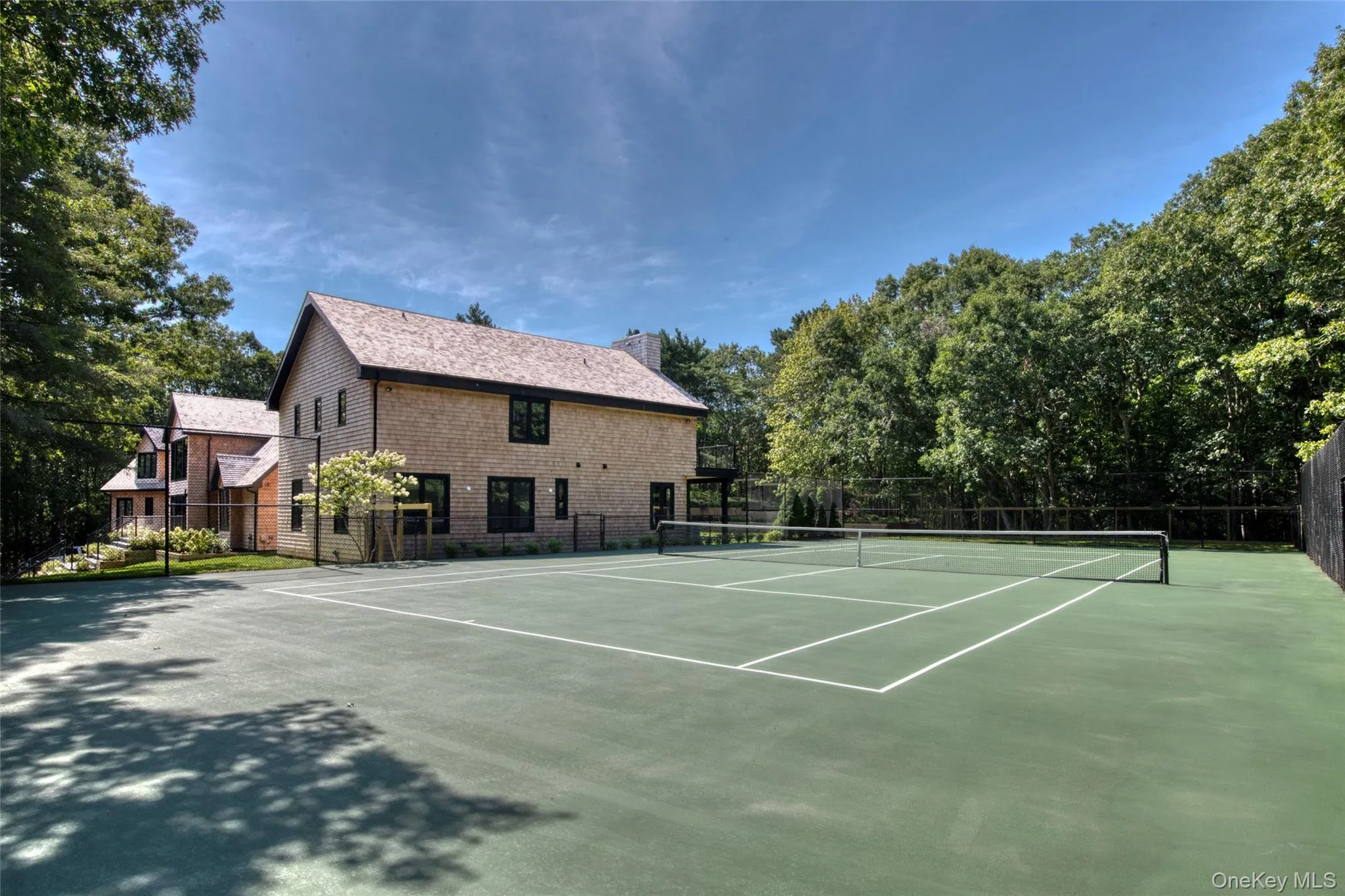 Full-size tennis court with a perimeter fence, adjacent to a residence with shingle siding and dark-trimmed windows Full-size tennis court with a perimeter fence, adjacent to a residence with shingle siding and dark-trimmed windows