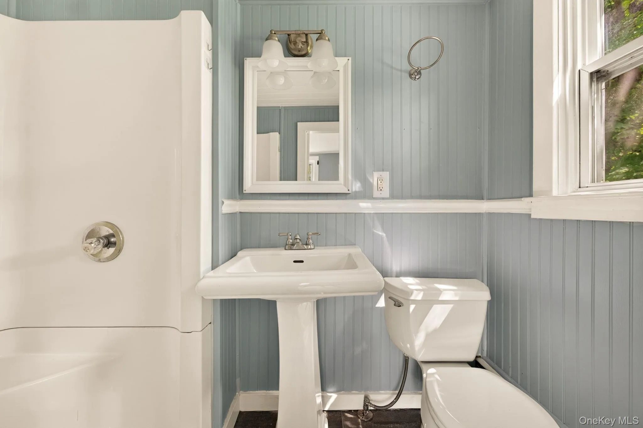 Compact bathroom featuring a pedestal sink with chrome fixtures, white porcelain toilet, vertical shiplap wall paneling in soft gray, framed mirror with white trim, and natural light from a window Compact bathroom featuring a pedestal sink with chrome fixtures, white porcelain toilet, vertical shiplap wall paneling in soft gray, framed mirror with white trim, and natural light from a window