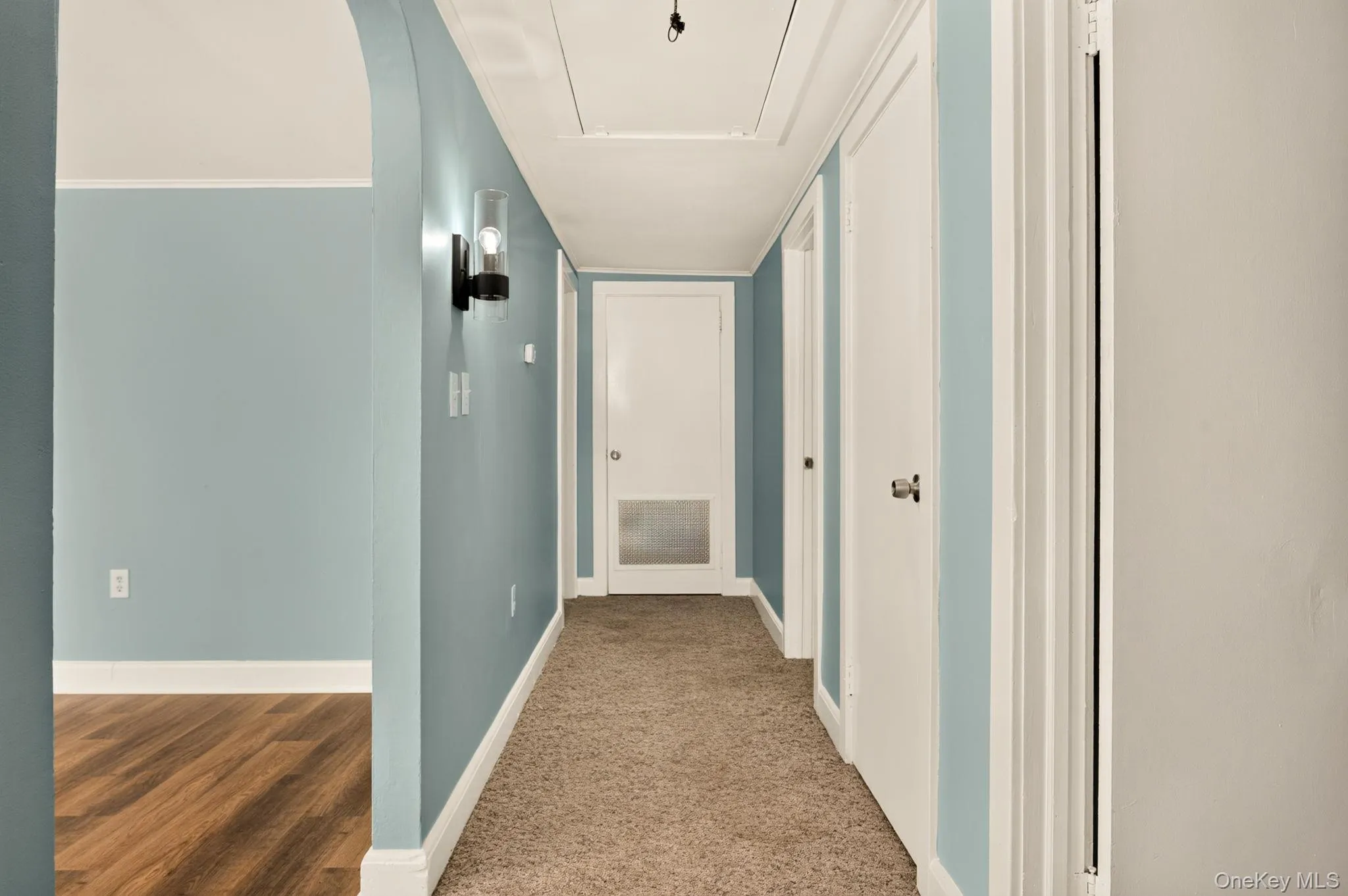 Hallway with textured carpeting, light blue walls, white trim, and a contemporary wall sconce Hallway with textured carpeting, light blue walls, white trim, and a contemporary wall sconce