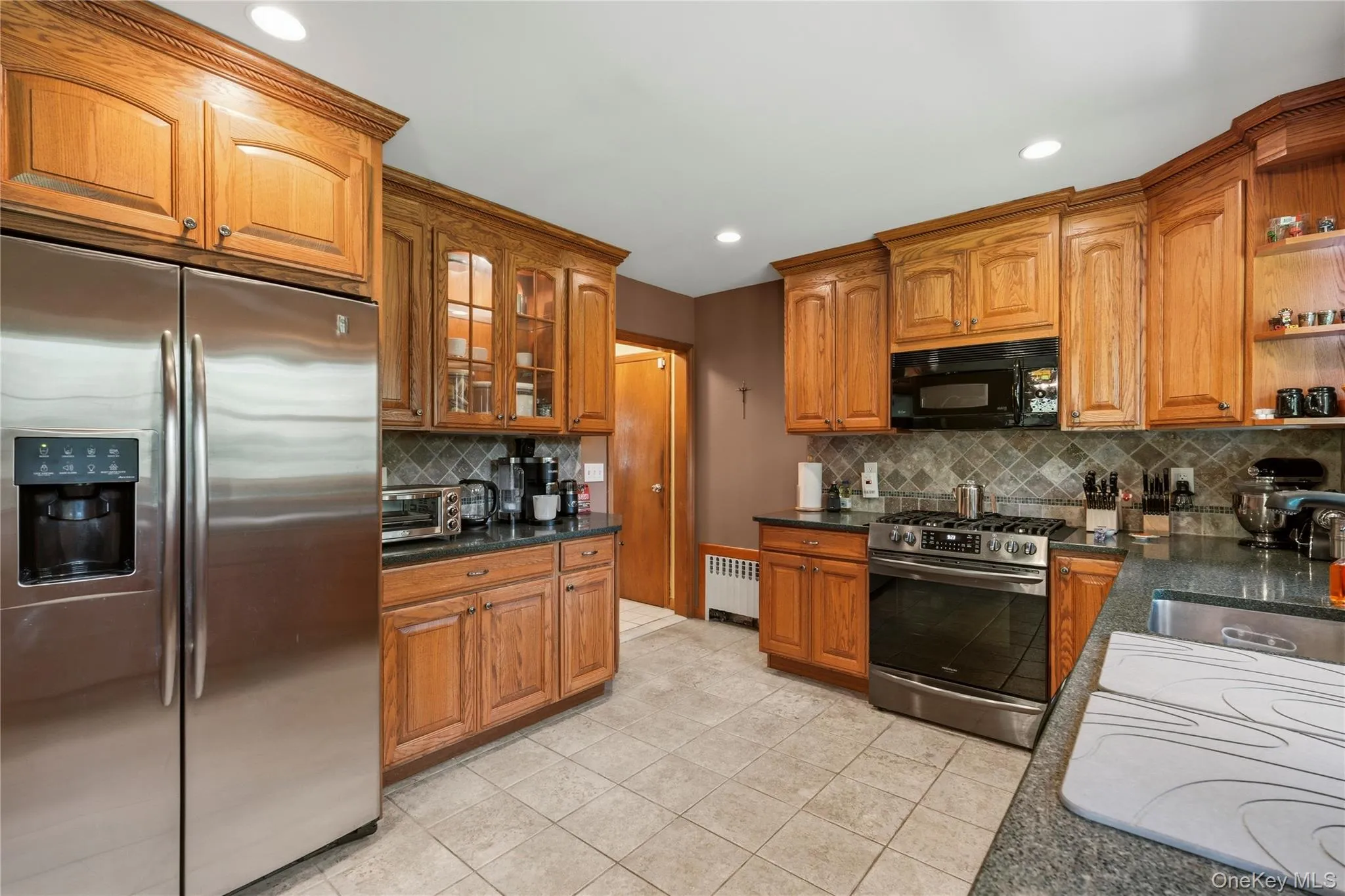 Kitchen featuring wood cabinetry, stainless steel appliances, dark countertops, a tiled backsplash, and recessed lighting Kitchen featuring wood cabinetry, stainless steel appliances, dark countertops, a tiled backsplash, and recessed lighting