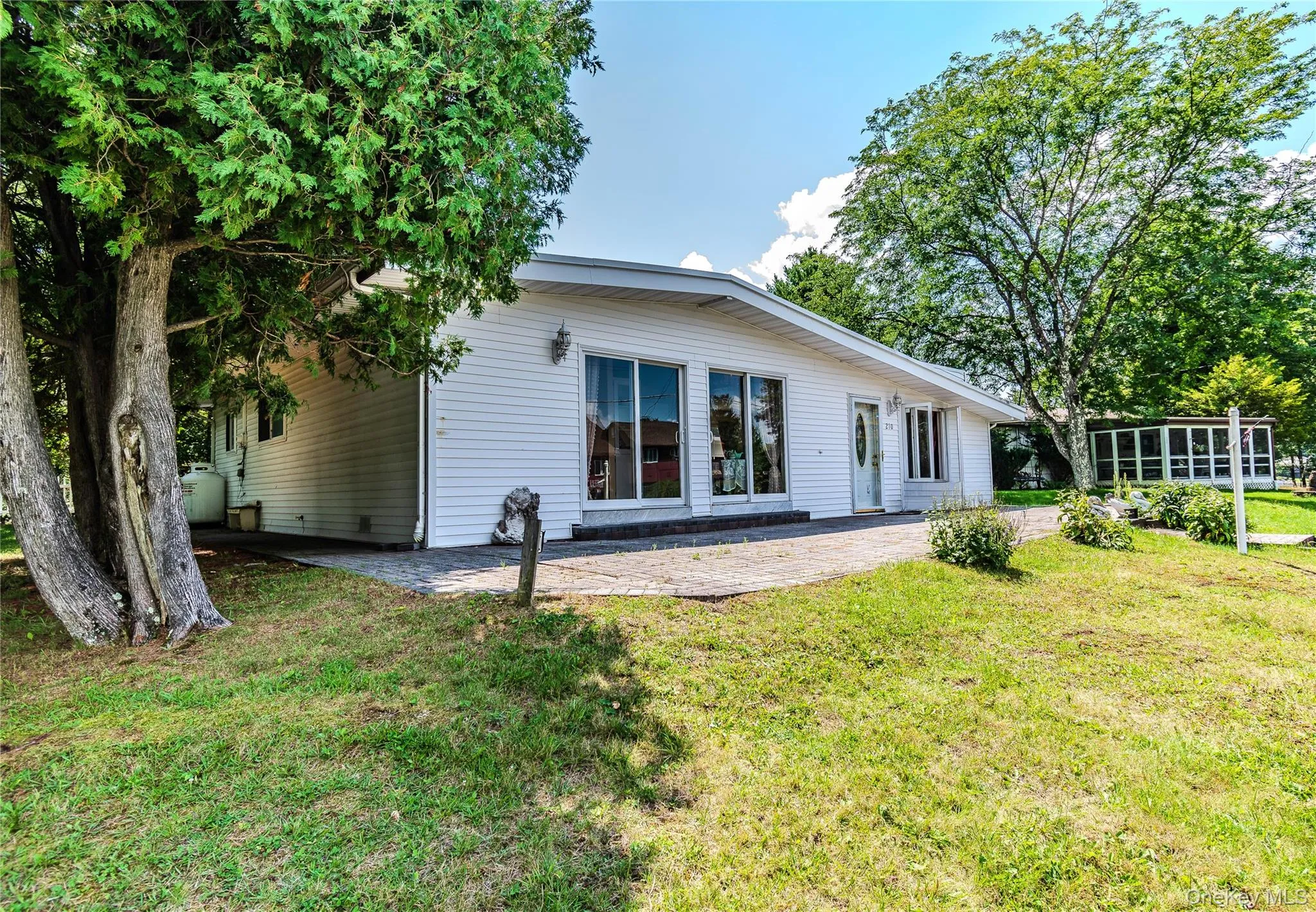 White siding exterior with multiple full-height windows, a paved patio, and mature trees White siding exterior with multiple full-height windows, a paved patio, and mature trees