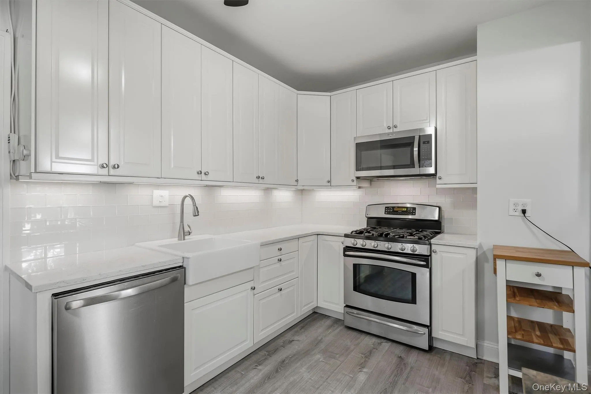 White cabinetry with brushed nickel hardware, stainless steel appliances, large farmhouse sink, subway tile backsplash, and wood-finish flooring White cabinetry with brushed nickel hardware, stainless steel appliances, large farmhouse sink, subway tile backsplash, and wood-finish flooring