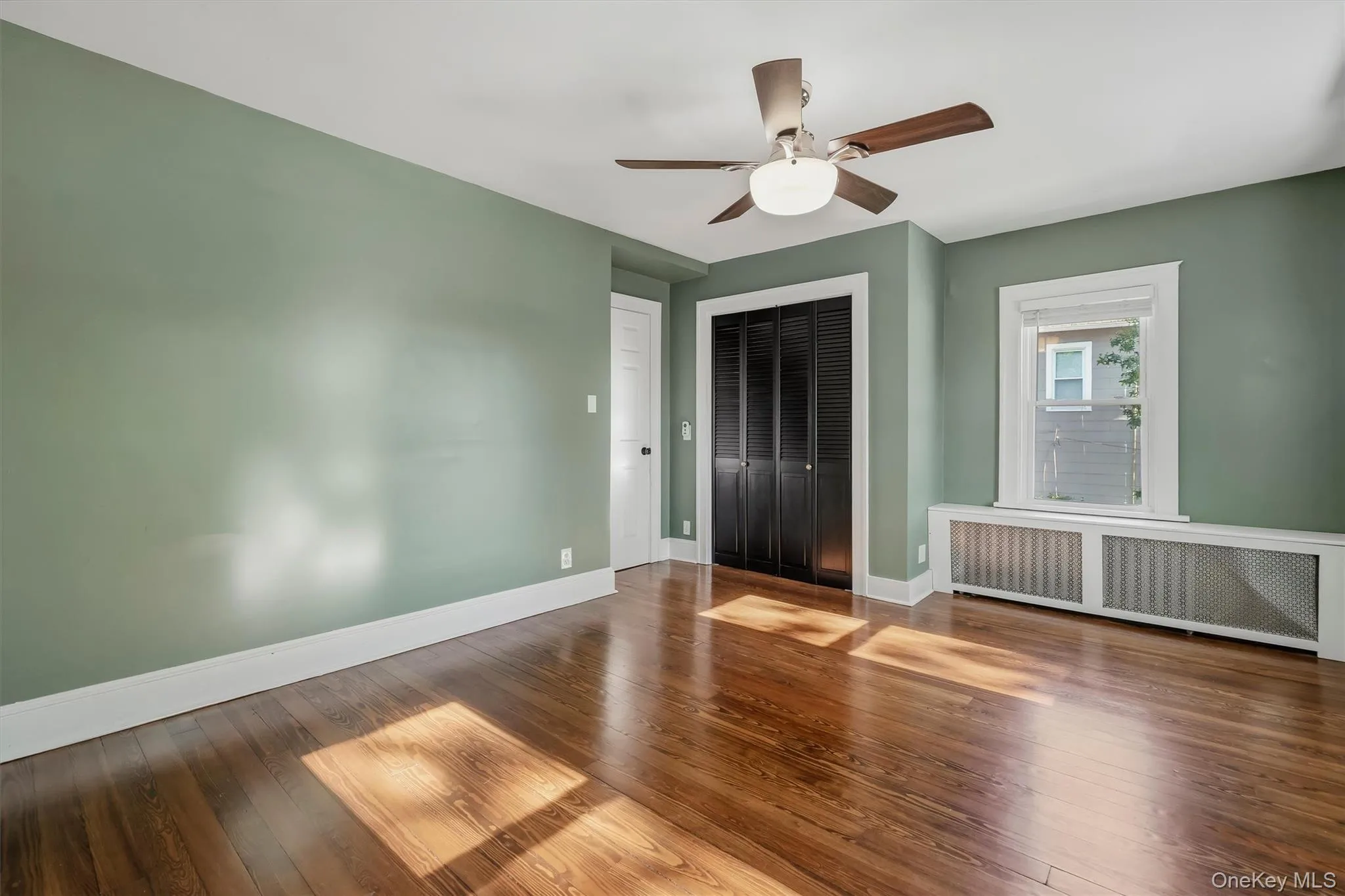 Wood-finish flooring, ceiling fan with integrated lighting, window with white trim, white door, and louvered bifold closet doors Wood-finish flooring, ceiling fan with integrated lighting, window with white trim, white door, and louvered bifold closet doors
