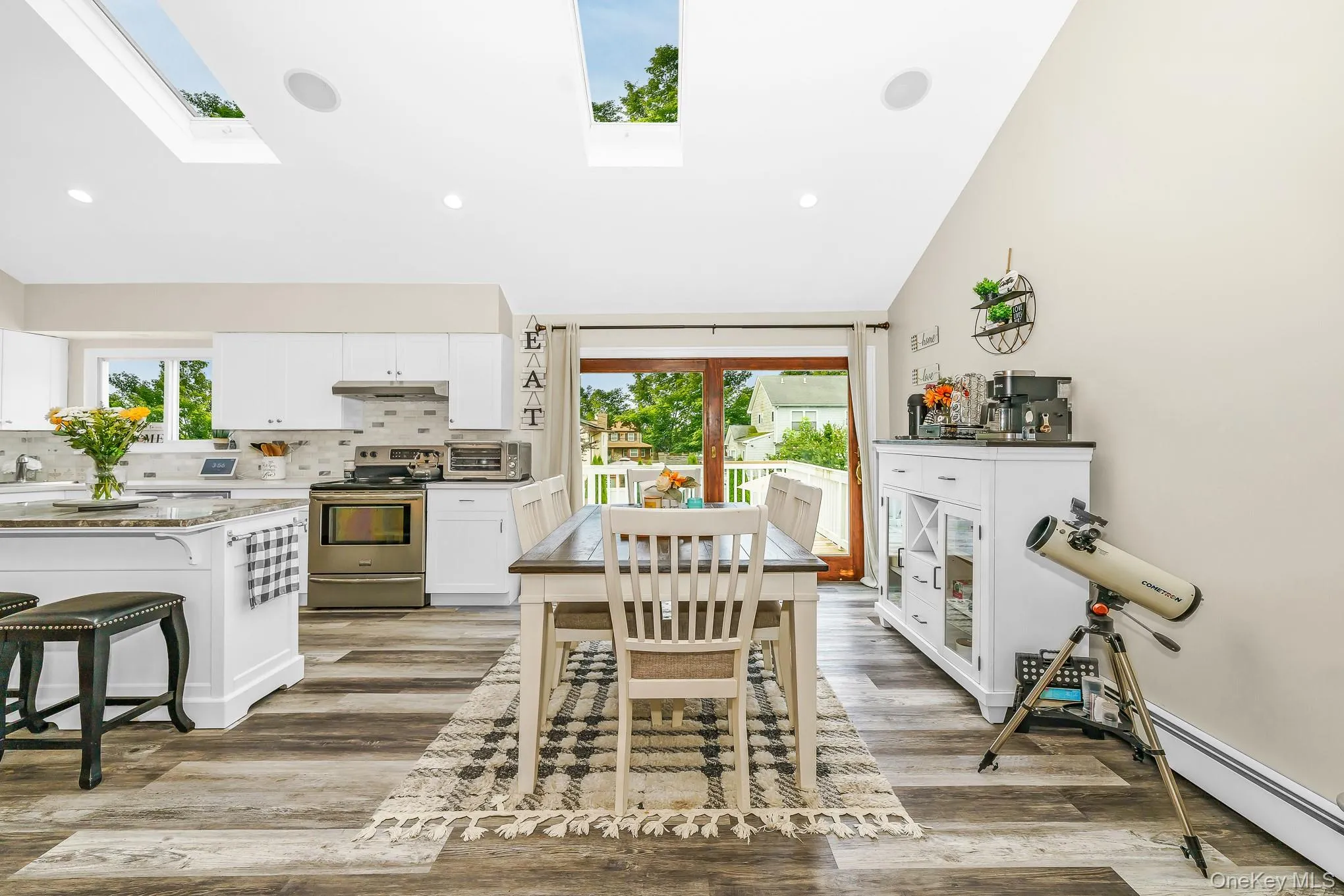 Open-concept kitchen and dining area featuring skylights, wood-finish flooring, white cabinetry, a kitchen island with seating, and stainless steel appliances Open-concept kitchen and dining area featuring skylights, wood-finish flooring, white cabinetry, a kitchen island with seating, and stainless steel appliances