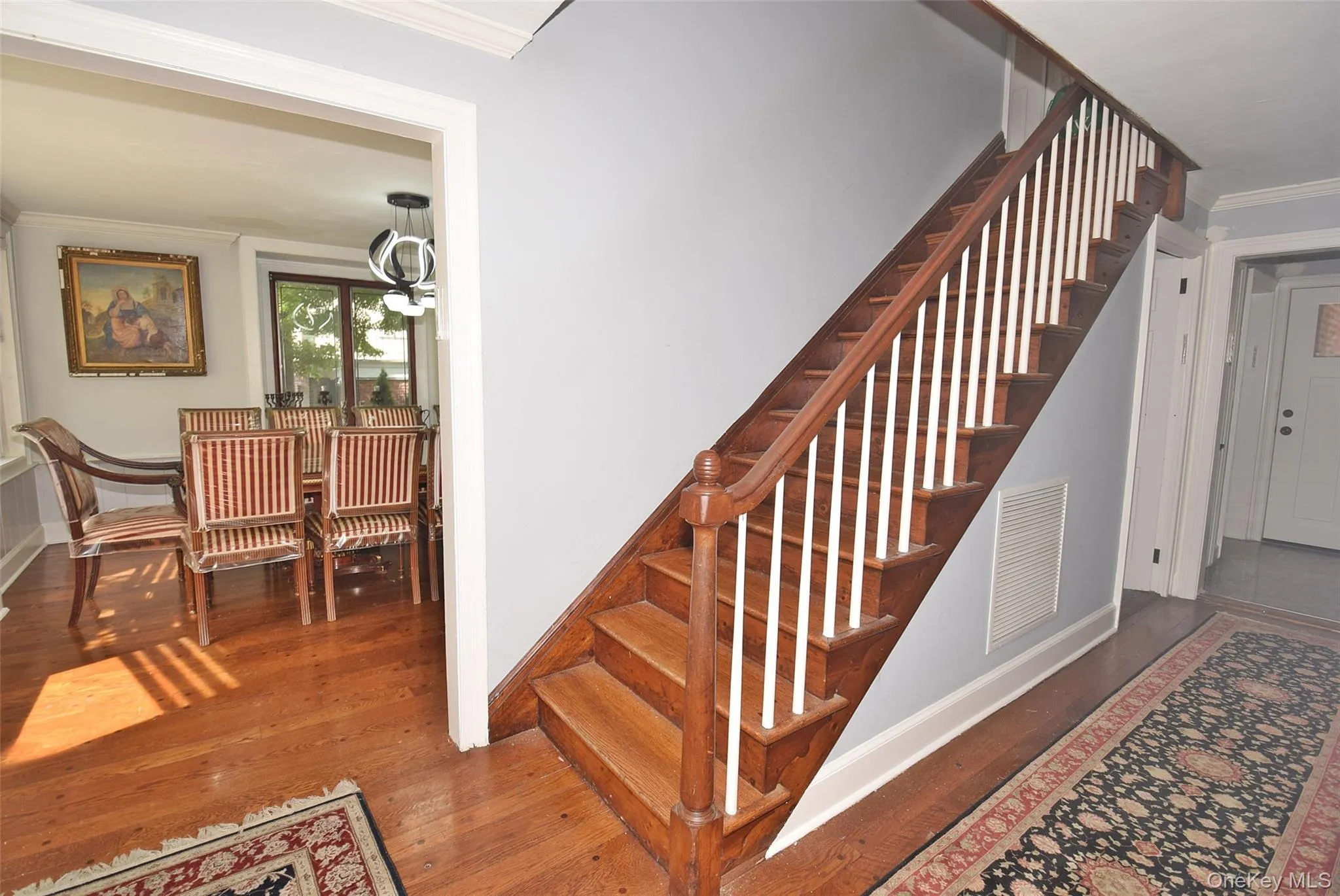 This image shows a home entryway with hardwood floors, a wooden staircase leading upstairs, a carpet runner down the hallway, and open doorways connecting to a dining area on the left and a living room on the right. This image shows a home entryway with hardwood floors, a wooden staircase leading upstairs, a carpet runner down the hallway, and open doorways connecting to a dining area on the left and a living room on the right.