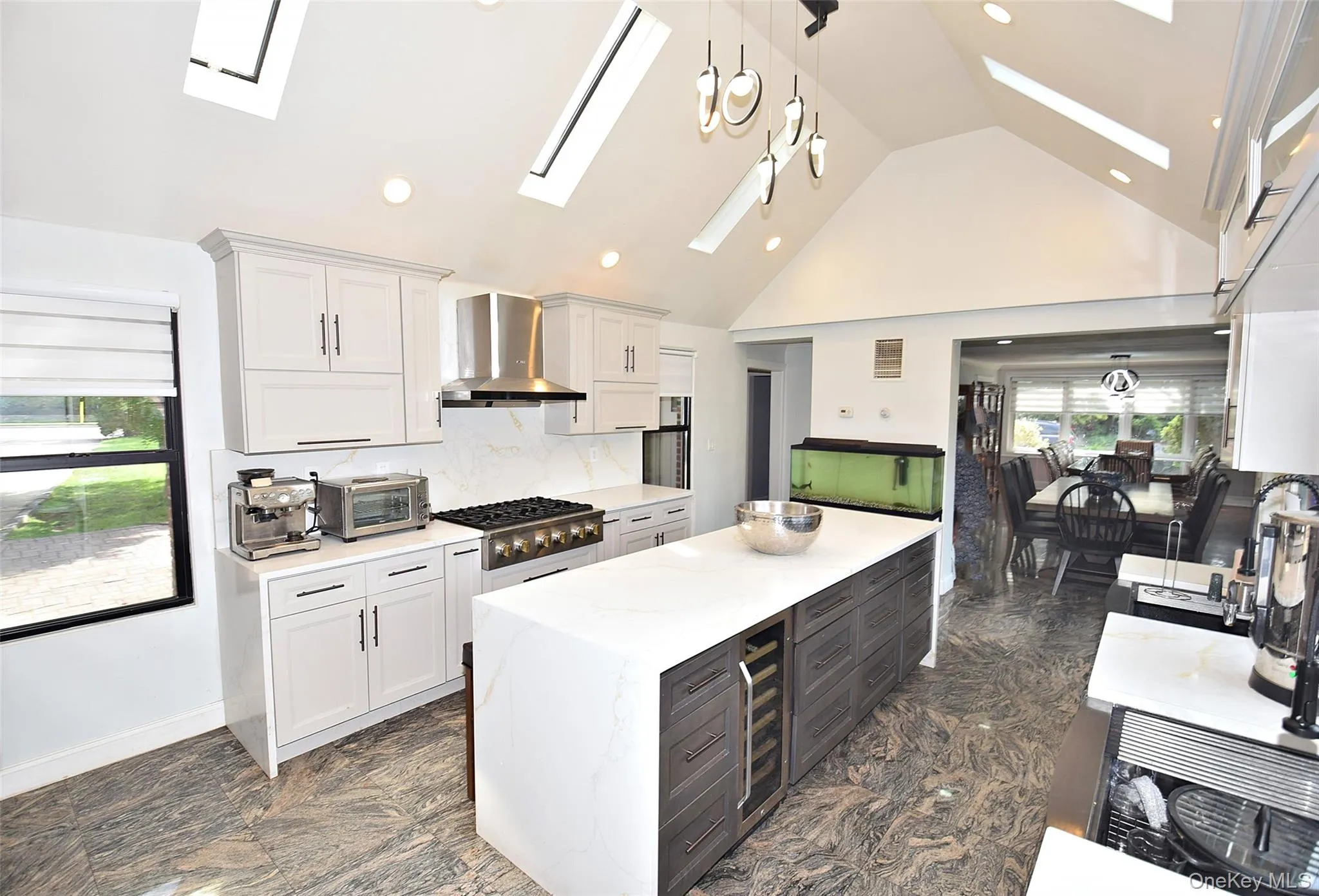 Vaulted ceiling with multiple skylights, kitchen island with built-in beverage cooler, stainless steel gas range with hood, light-toned cabinetry, and extensive counter space Vaulted ceiling with multiple skylights, kitchen island with built-in beverage cooler, stainless steel gas range with hood, light-toned cabinetry, and extensive counter space
