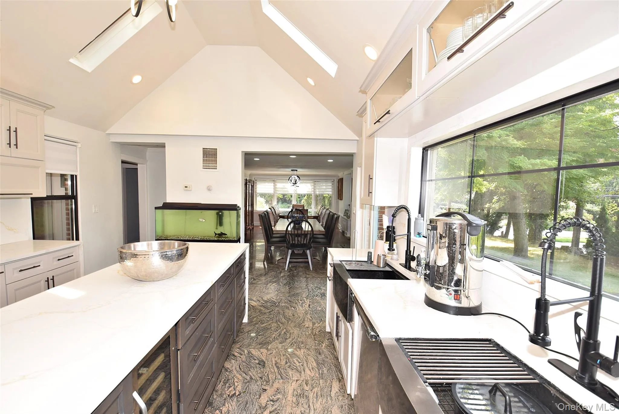 Vaulted ceiling with skylights, white cabinetry, integrated range hood, built-in wine refrigerator, and a large window above the kitchen 2 sinks. Vaulted ceiling with skylights, white cabinetry, integrated range hood, built-in wine refrigerator, and a large window above the kitchen 2 sinks.