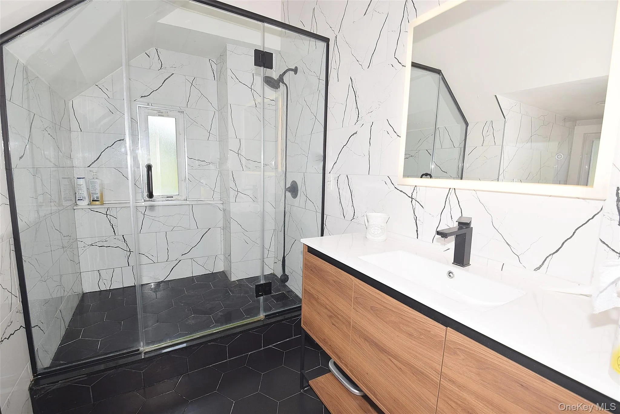 Frameless glass shower enclosure with black trim, a wood-finish vanity with an integrated sink, black hexagonal floor tiles, and a wall-mounted lighted mirror Frameless glass shower enclosure with black trim, a wood-finish vanity with an integrated sink, black hexagonal floor tiles, and a wall-mounted lighted mirror