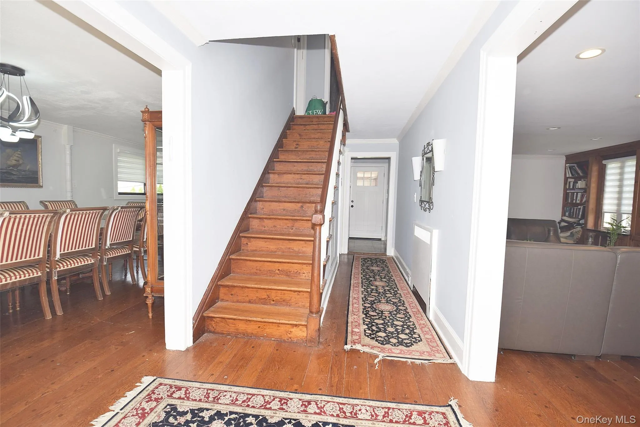 This image shows a home entryway with hardwood floors, a wooden staircase leading upstairs, a carpet runner down the hallway, and open doorways connecting to a dining area on the left and a living room on the right. This image shows a home entryway with hardwood floors, a wooden staircase leading upstairs, a carpet runner down the hallway, and open doorways connecting to a dining area on the left and a living room on the right.