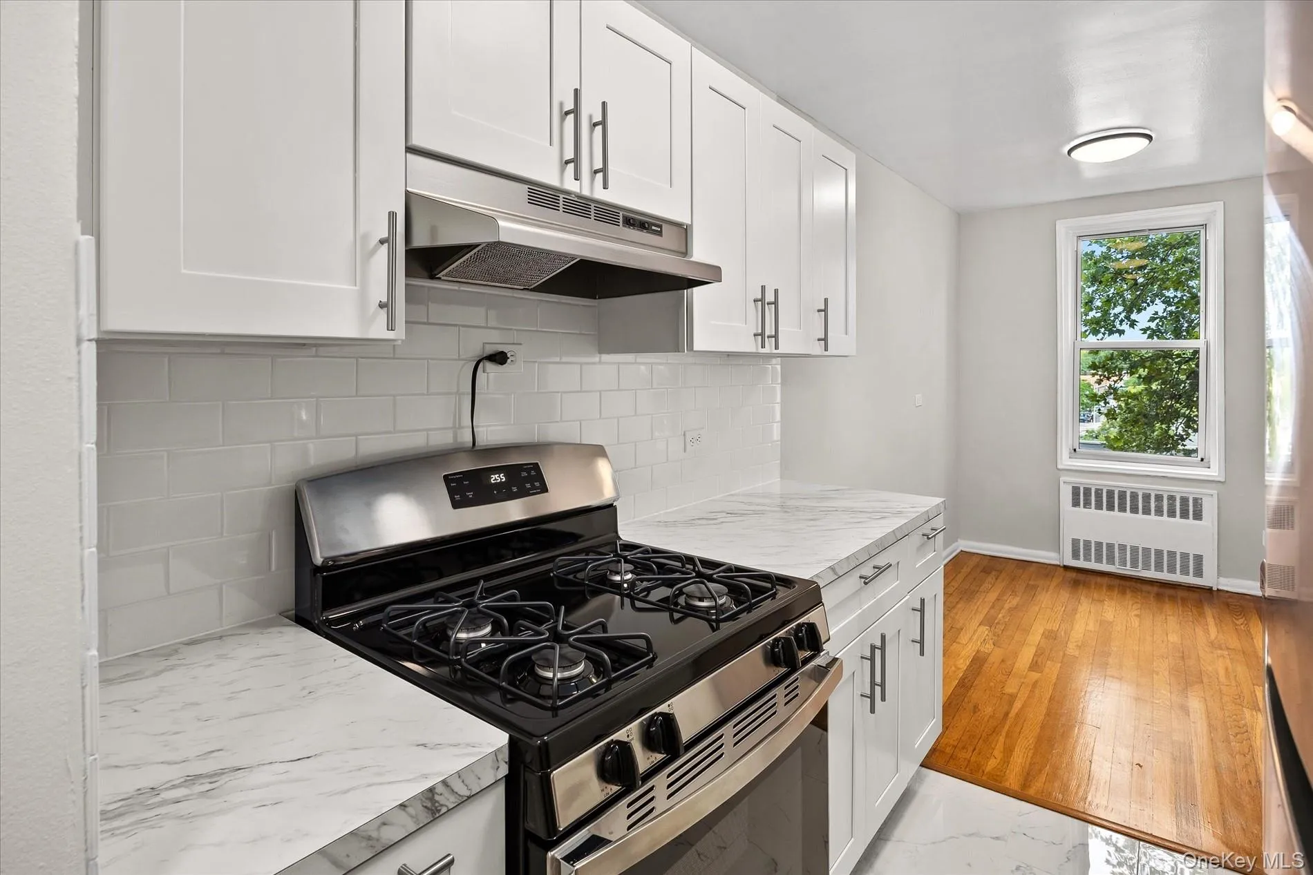 Kitchen featuring white shaker cabinetry, white subway tile backsplash, stainless steel range with overhead ventilation, wood-finish flooring, and a large window Kitchen featuring white shaker cabinetry, white subway tile backsplash, stainless steel range with overhead ventilation, wood-finish flooring, and a large window