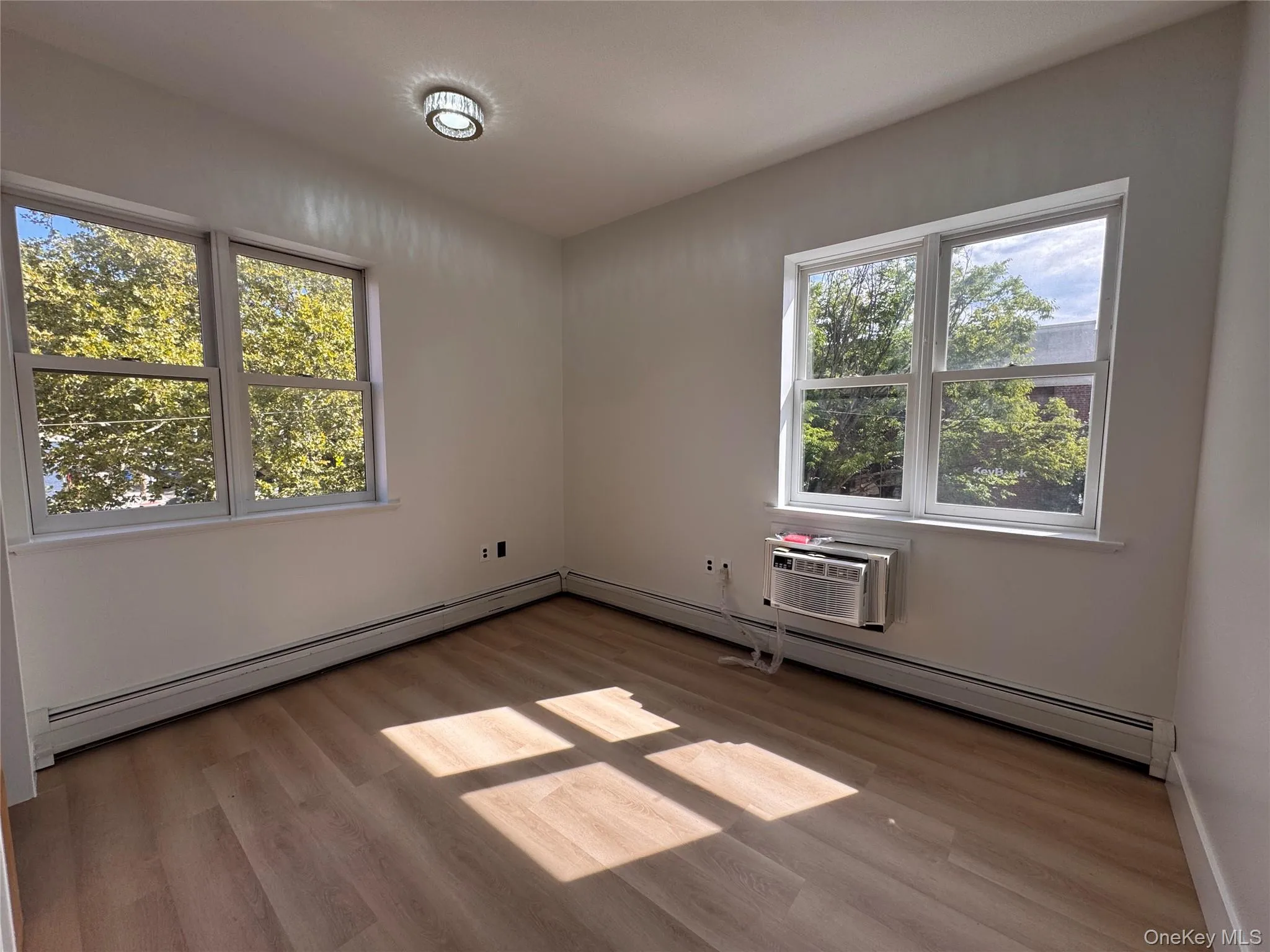 Wood-finish flooring, multiple windows, window-mounted air conditioning unit, ceiling-mounted light fixture, and white wall finishes Wood-finish flooring, multiple windows, window-mounted air conditioning unit, ceiling-mounted light fixture, and white wall finishes