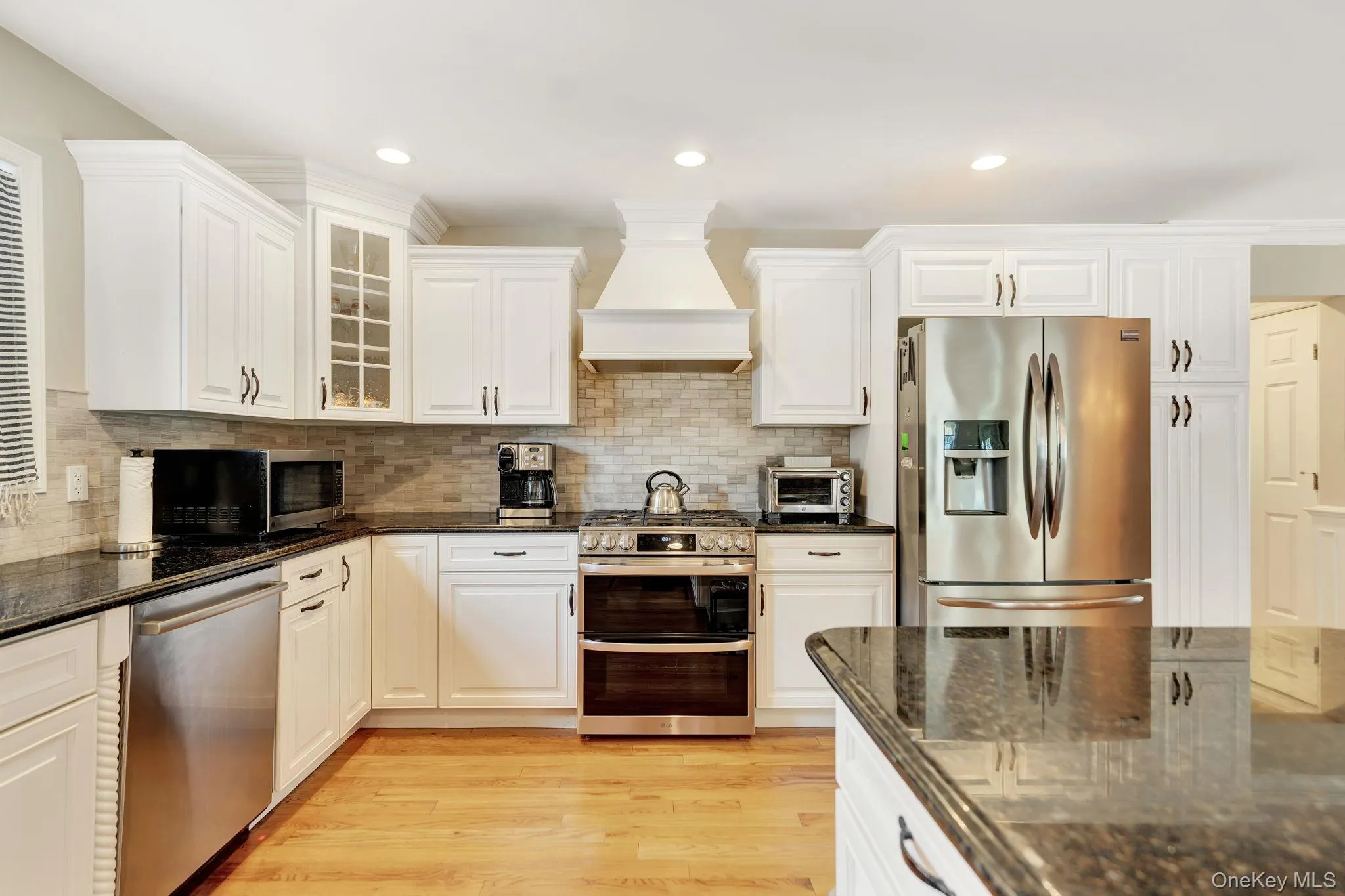 White cabinetry with dark hardware, stainless steel appliances, wood-finish flooring, granite countertops, and a tile backsplash White cabinetry with dark hardware, stainless steel appliances, wood-finish flooring, granite countertops, and a tile backsplash