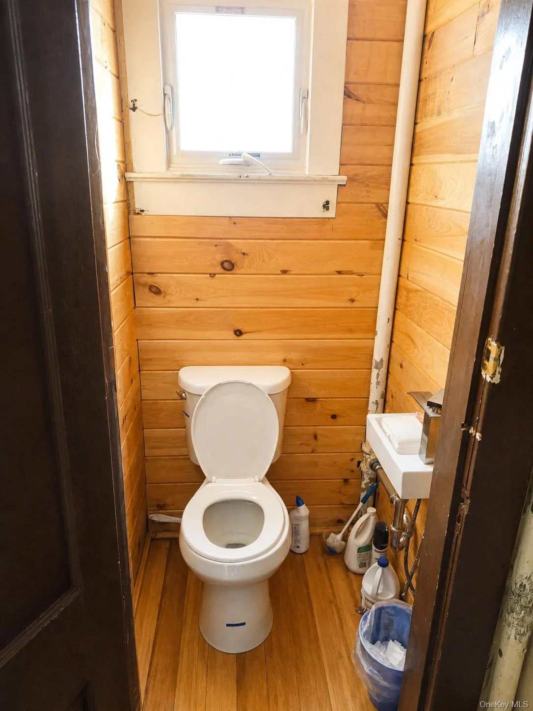 Wood-paneled walls with a casement window, wood-finish flooring, and a white toilet Wood-paneled walls with a casement window, wood-finish flooring, and a white toilet