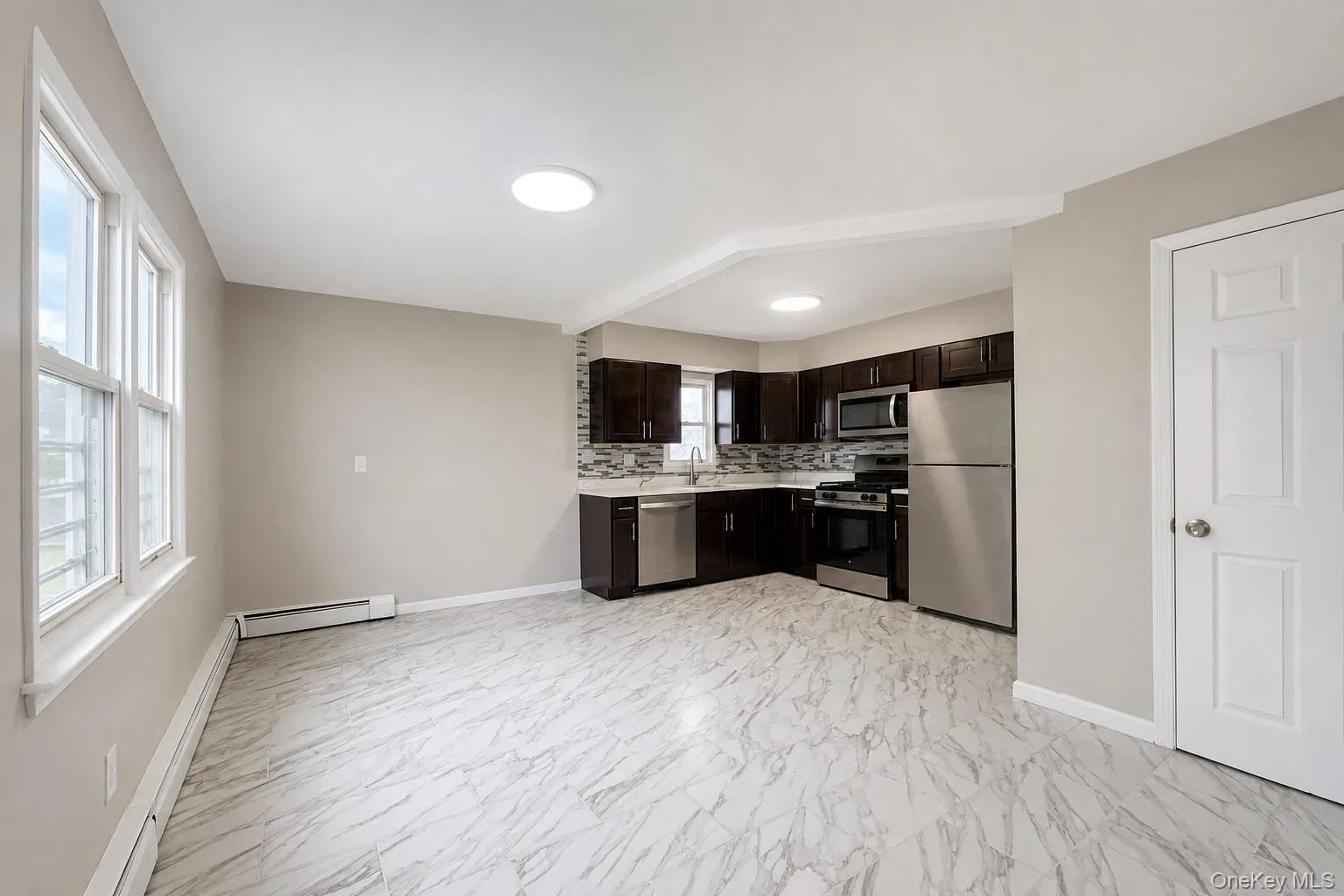 Open-concept kitchen and living area featuring dark wood-finish cabinetry, stainless steel appliances, and a mosaic tile backsplash Open-concept kitchen and living area featuring dark wood-finish cabinetry, stainless steel appliances, and a mosaic tile backsplash