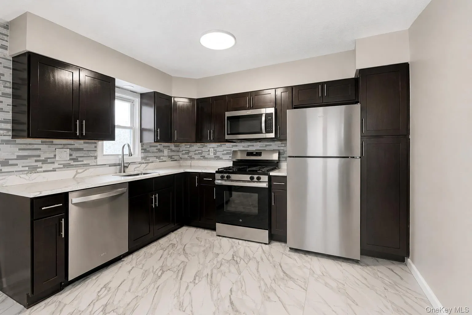 Open-concept kitchen and living area featuring dark wood-finish cabinetry, stainless steel appliances, and a mosaic tile backsplash Open-concept kitchen and living area featuring dark wood-finish cabinetry, stainless steel appliances, and a mosaic tile backsplash