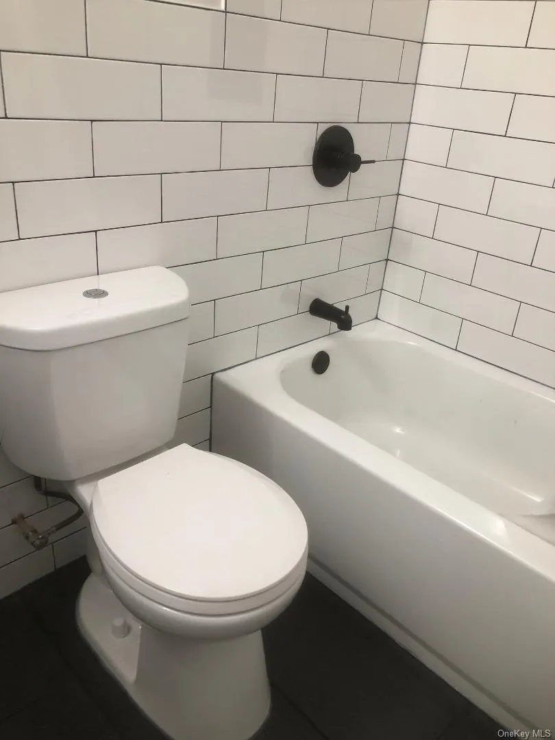 Bathroom featuring a shower/tub combination, white subway tile walls, a standard white toilet, a white bathtub, dark floor tiling, and matte black fixtures Bathroom featuring a shower/tub combination, white subway tile walls, a standard white toilet, a white bathtub, dark floor tiling, and matte black fixtures
