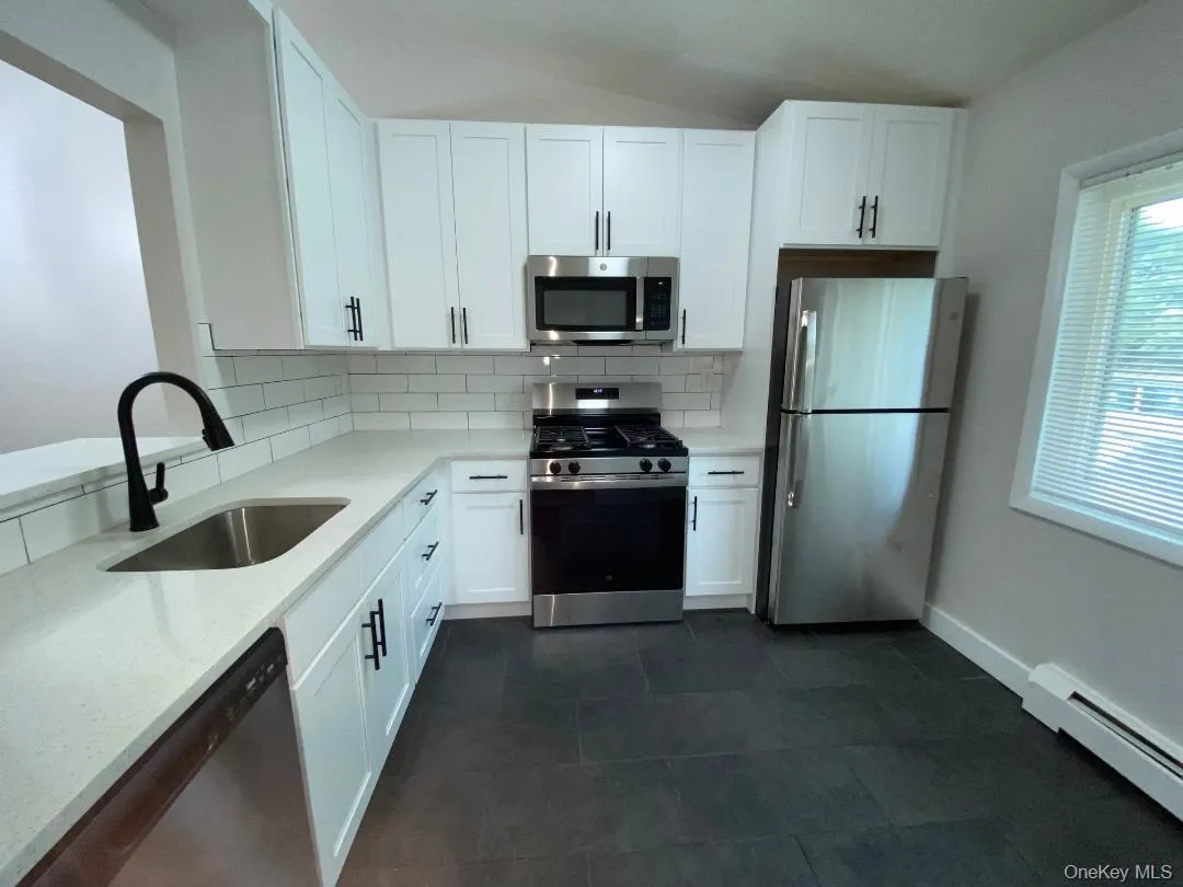 White shaker cabinetry with matte black hardware, stainless steel appliances, white subway tile backsplash, light-toned countertops, and dark tile flooring White shaker cabinetry with matte black hardware, stainless steel appliances, white subway tile backsplash, light-toned countertops, and dark tile flooring