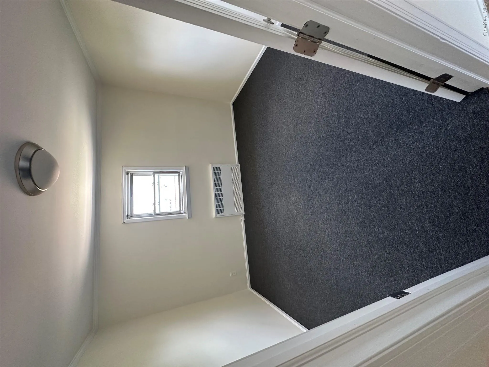 Dark gray carpeting, window with white trim, wall-mounted radiator, and flush-mount ceiling light Dark gray carpeting, window with white trim, wall-mounted radiator, and flush-mount ceiling light