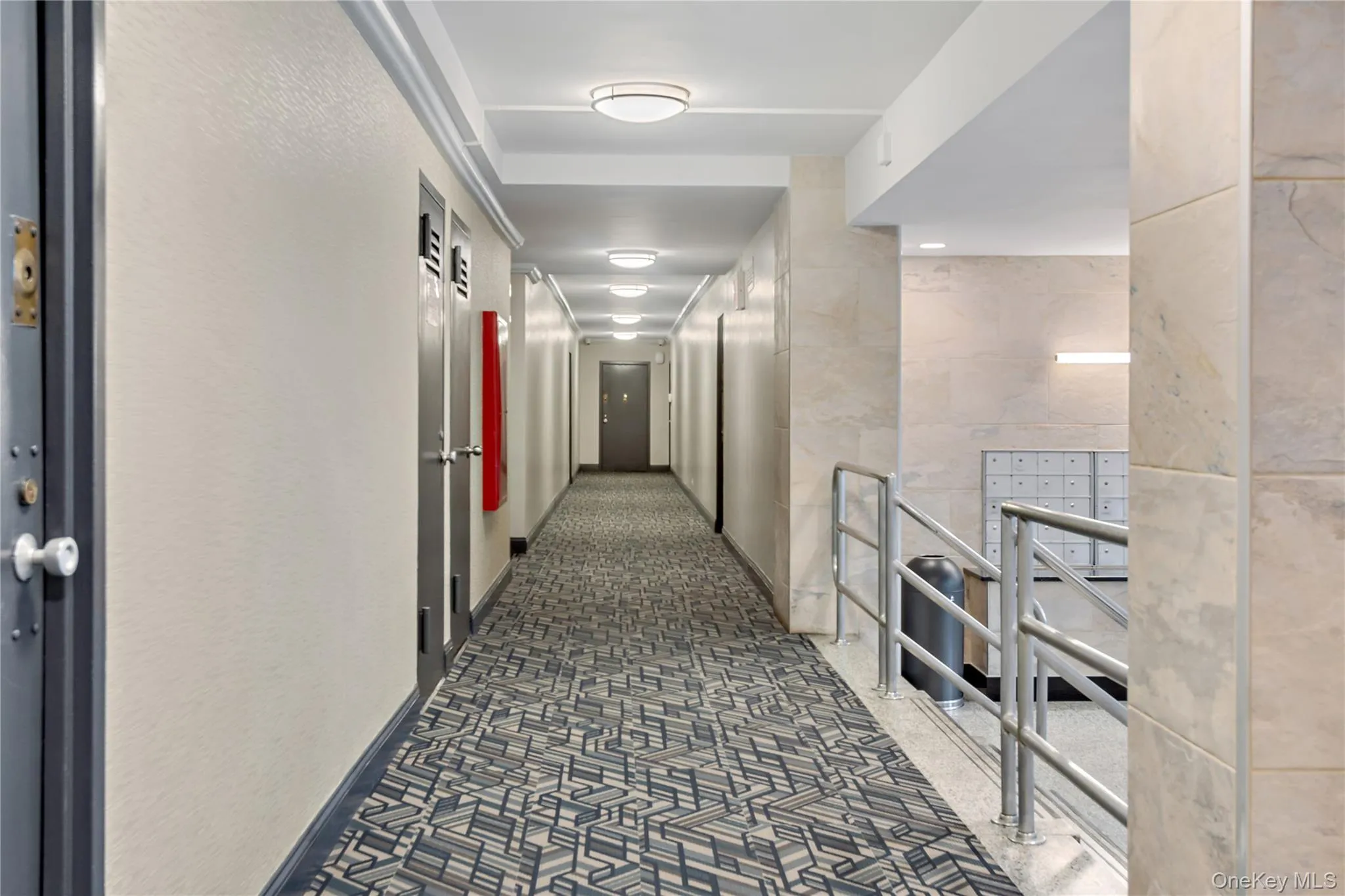 Well-lit corridor featuring recessed ceiling lighting, neutral wall finishes, geometric patterned flooring, and built-in mailbox fixtures along the passage Well-lit corridor featuring recessed ceiling lighting, neutral wall finishes, geometric patterned flooring, and built-in mailbox fixtures along the passage