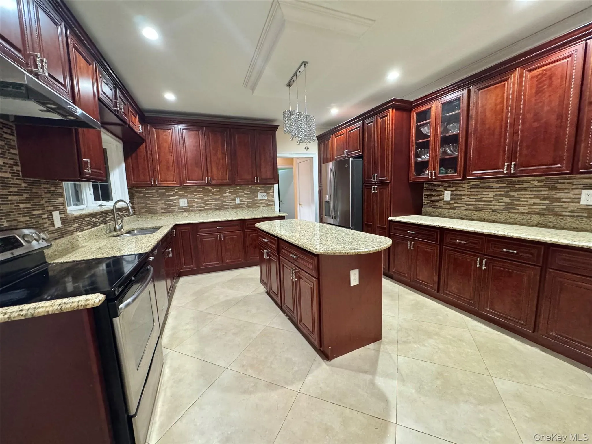 Expansive kitchen featuring wood cabinetry, light-toned countertops, a central island, stainless steel appliances, and a mosaic tile backsplash Expansive kitchen featuring wood cabinetry, light-toned countertops, a central island, stainless steel appliances, and a mosaic tile backsplash