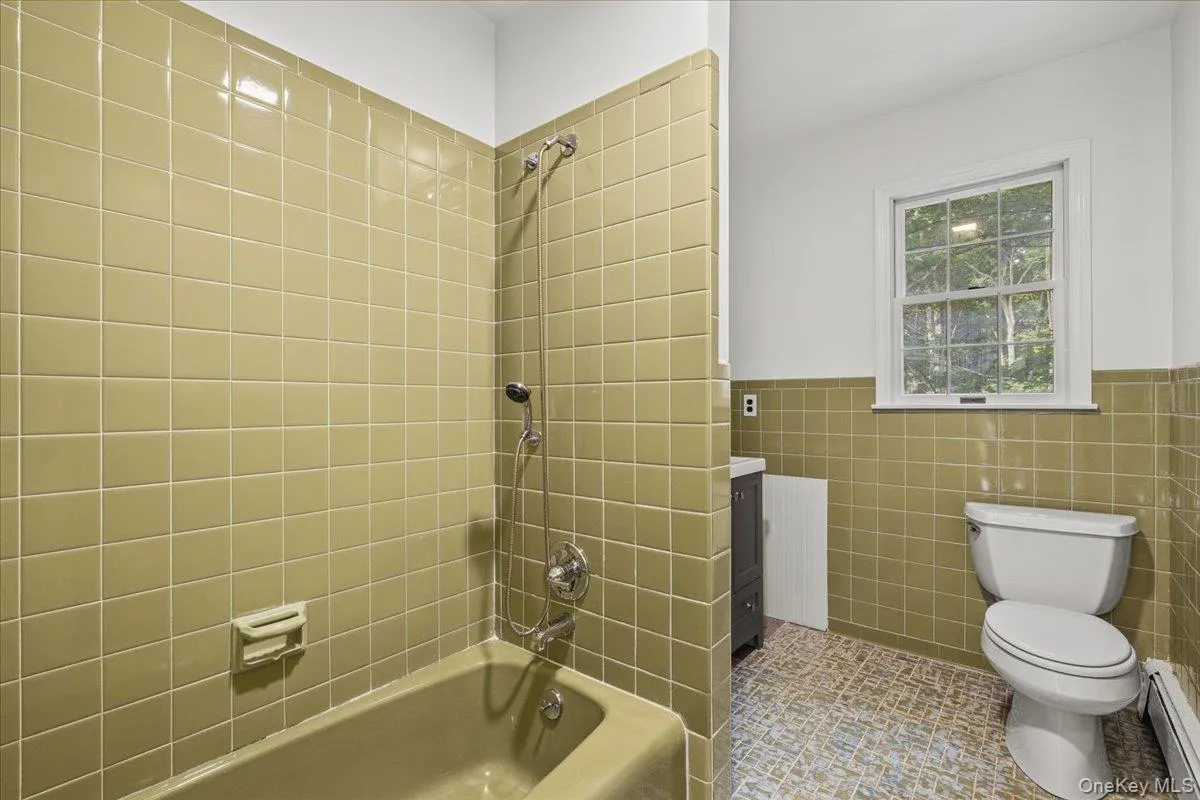 Bathroom featuring ceramic wall tiling, a built-in tub with a shower fixture, a window, and a toilet Bathroom featuring ceramic wall tiling, a built-in tub with a shower fixture, a window, and a toilet