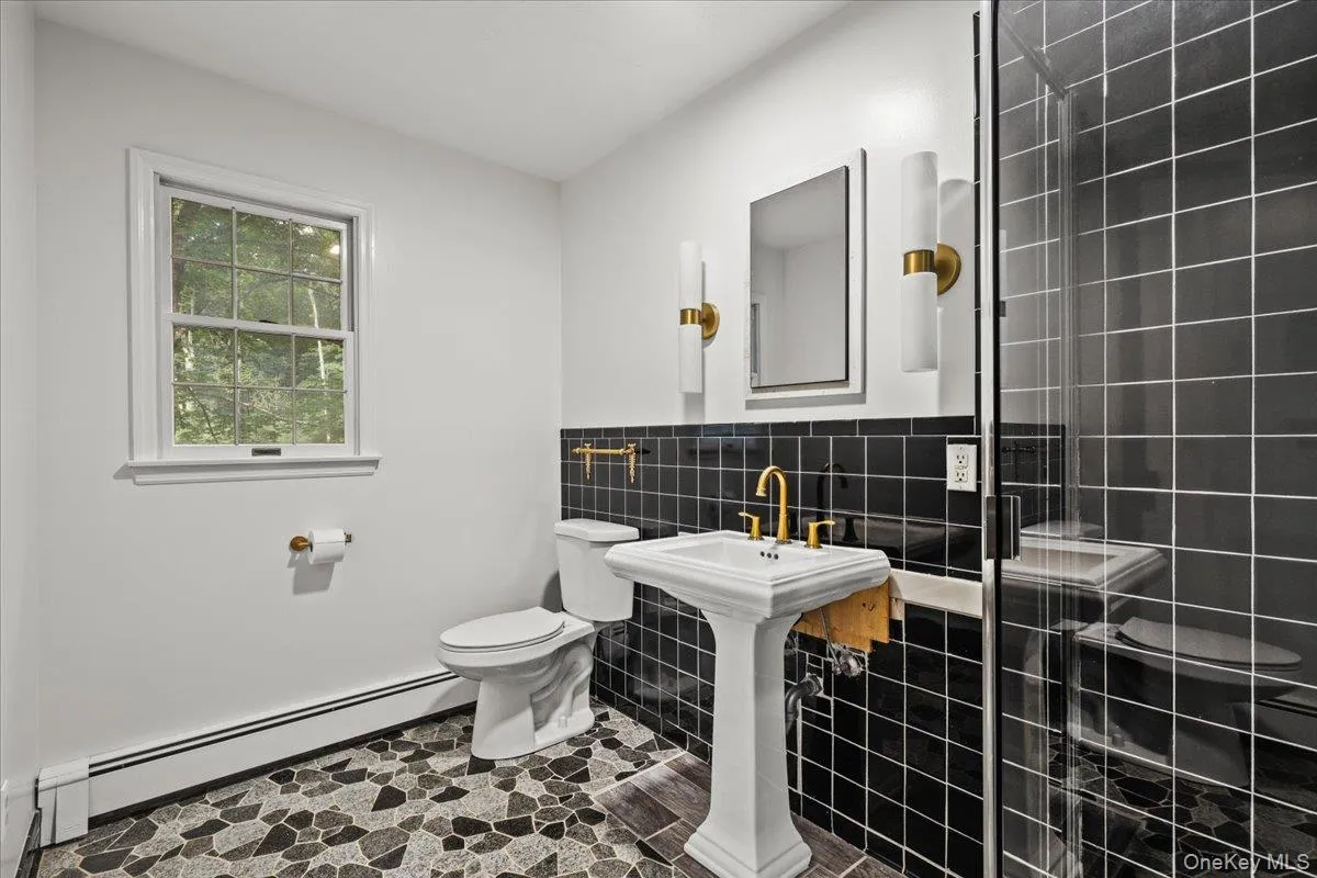 Pedestal sink with brass-finish faucet, black tiled walls, glass-enclosed shower, and multi-tone stone flooring Pedestal sink with brass-finish faucet, black tiled walls, glass-enclosed shower, and multi-tone stone flooring