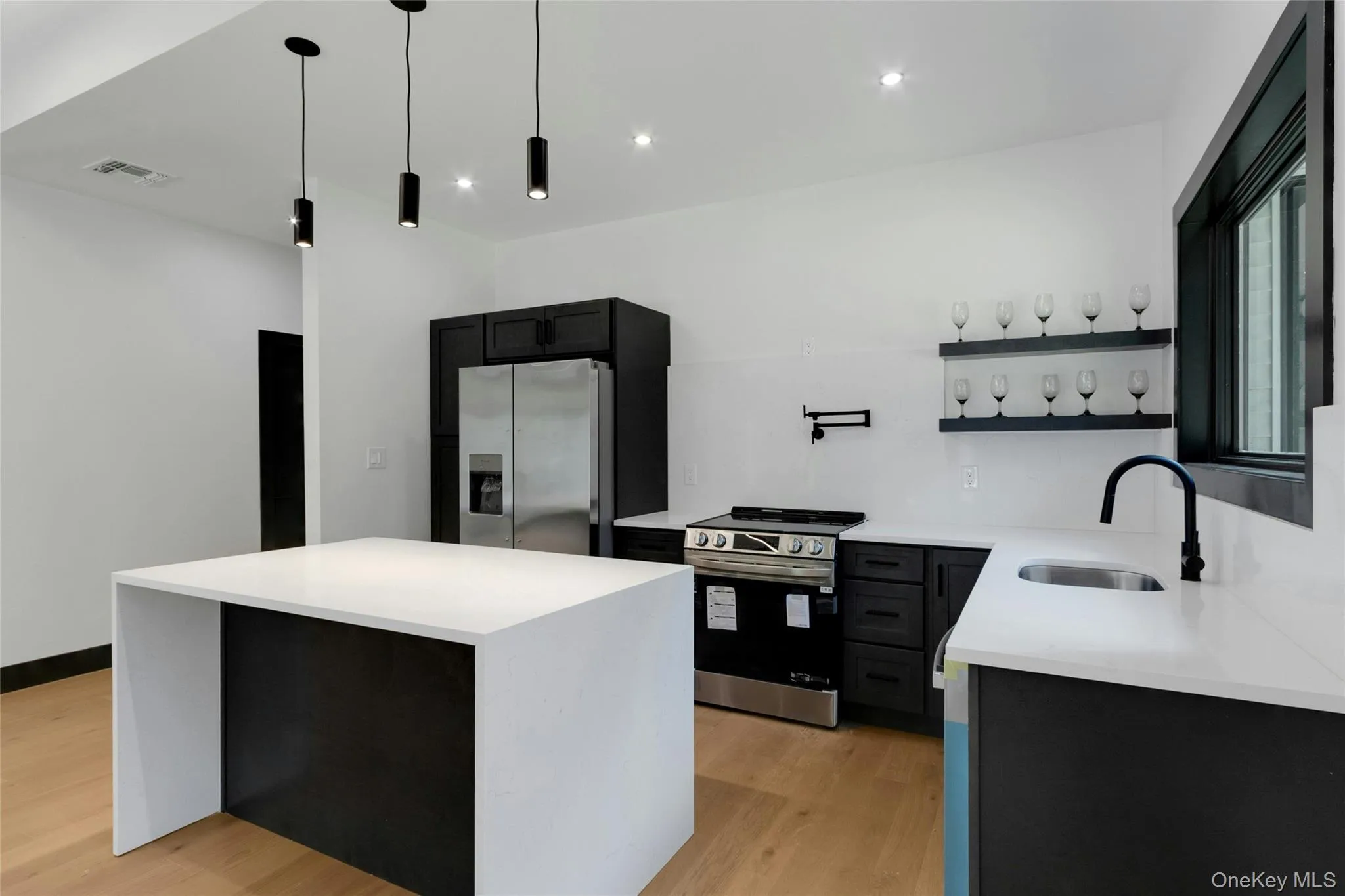 Contemporary kitchen featuring a white center island with a waterfall edge, black cabinetry, white countertops, and stainless steel appliances Contemporary kitchen featuring a white center island with a waterfall edge, black cabinetry, white countertops, and stainless steel appliances