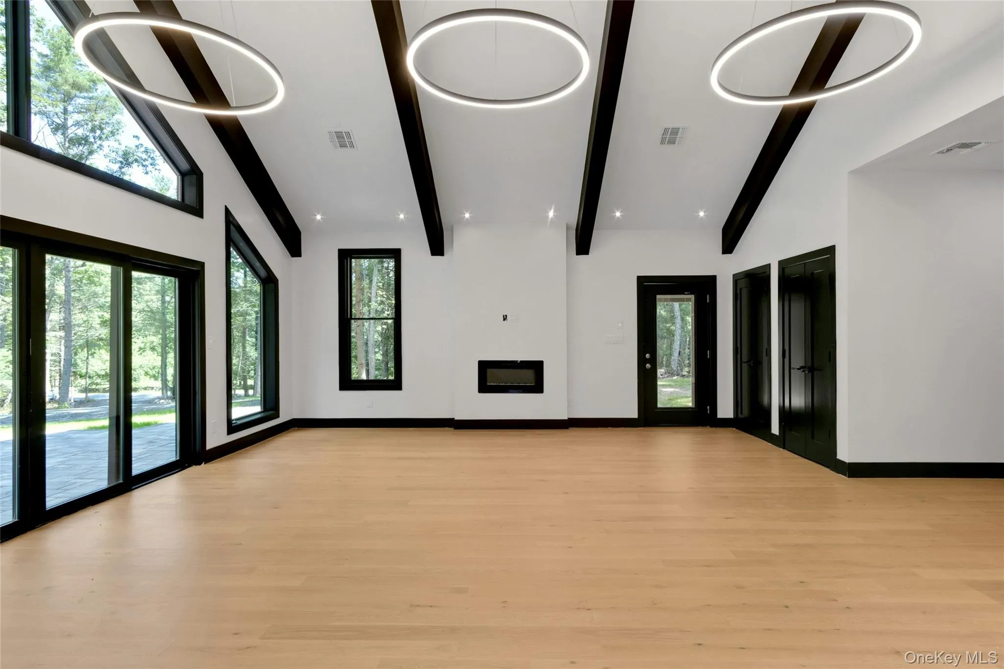 Vaulted ceiling with exposed beams, light wood-finish flooring, linear electric fireplace, expansive glass sliding doors, and contemporary circular pendant lighting Vaulted ceiling with exposed beams, light wood-finish flooring, linear electric fireplace, expansive glass sliding doors, and contemporary circular pendant lighting