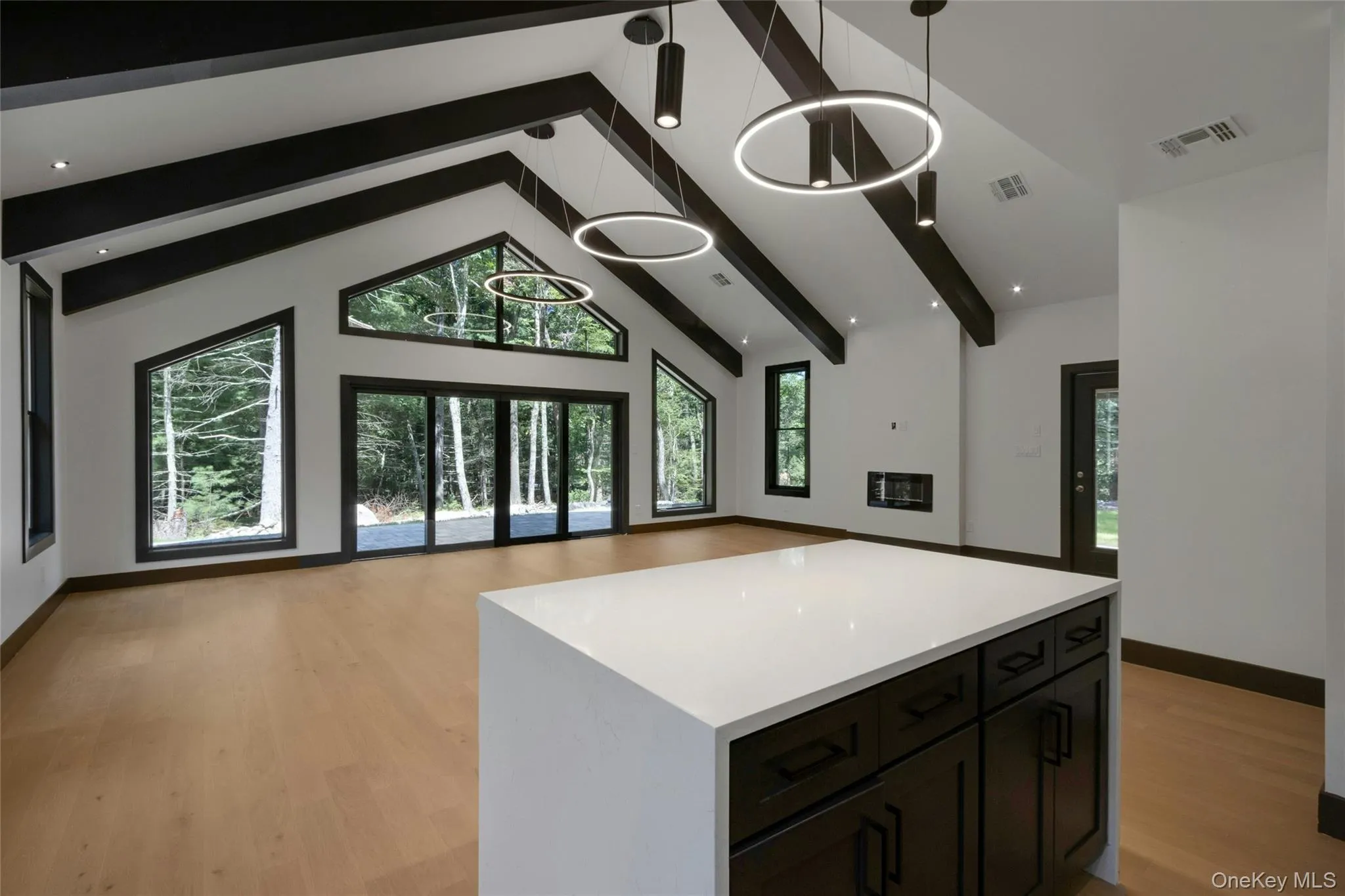 Vaulted ceiling with exposed beams, wood-finish flooring, built-in electric fireplace, contemporary pendant lighting, and a kitchen island with a white countertop and dark wood cabinetry Vaulted ceiling with exposed beams, wood-finish flooring, built-in electric fireplace, contemporary pendant lighting, and a kitchen island with a white countertop and dark wood cabinetry